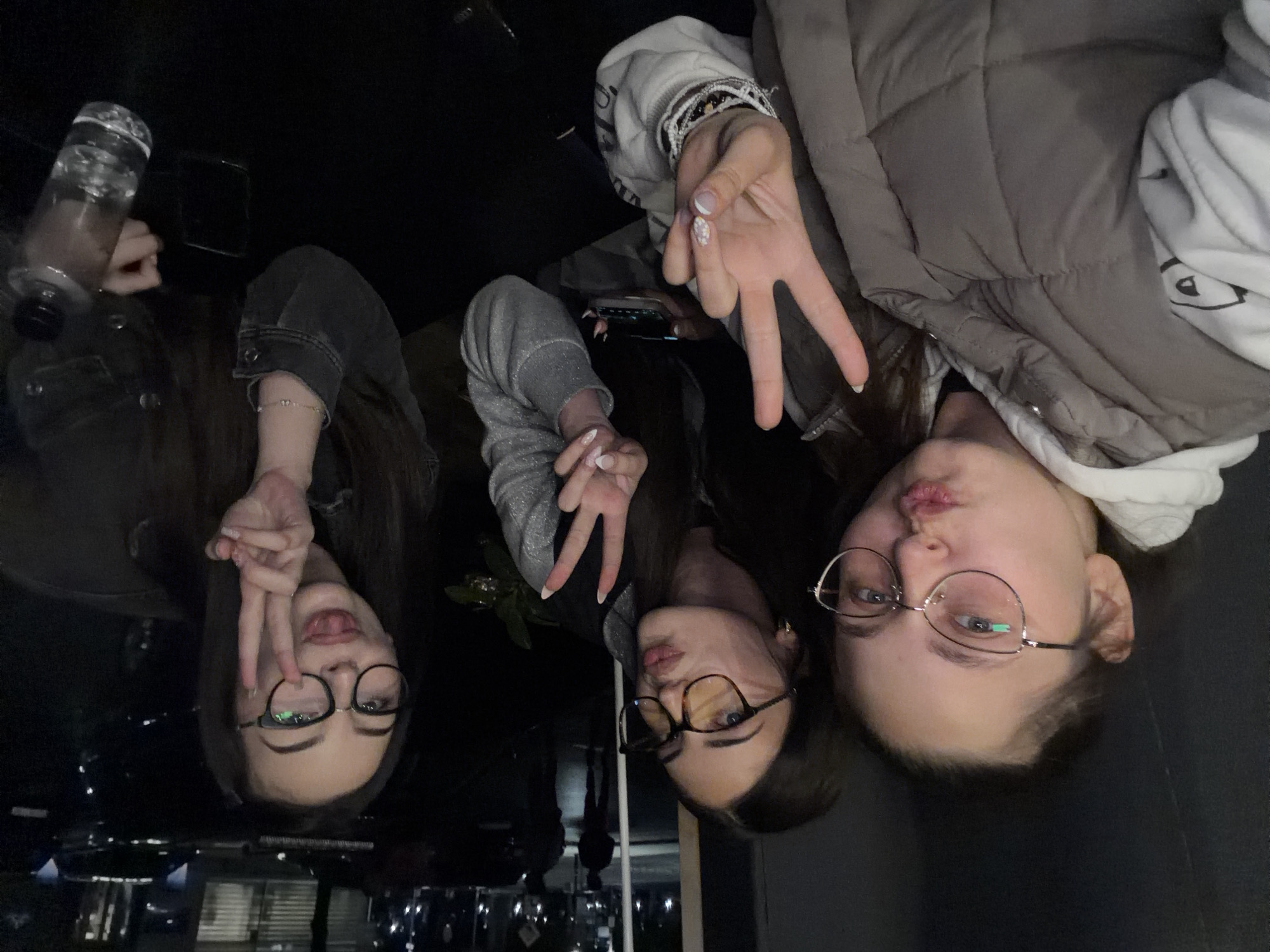 Three young women are posed close together, making peace sign hand gestures. The woman on the right wears a puffy gray vest over a white hooded sweatshirt. She has light skin, dark hair, wears glasses, and her lips are pursed. The middle woman wears a silver-gray shiny sweatshirt, and her hair is dark. She is wearing dark-framed glasses. The left woman wears a dark denim jacket and eyeglasses. She has dark hair and is sticking out her tongue. There is a bottle of water and a phone on a dark surface to the left. It is impossible to determine the location within Montana, Bulgaria from the visual information provided. The setting appears to be dimly lit indoors.