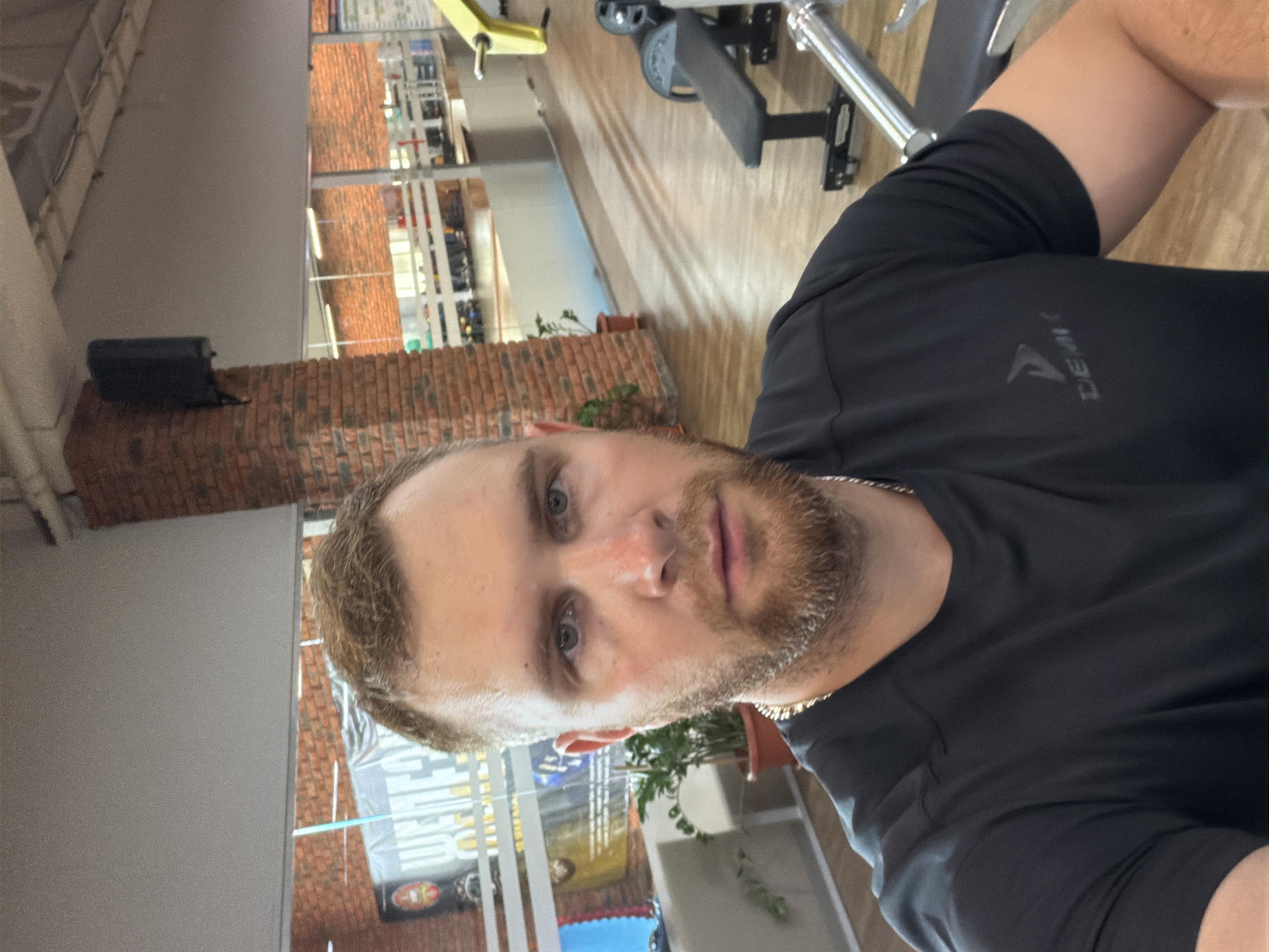 A man with light brown hair and a beard, wearing a black t-shirt and a silver chain, takes a selfie in a gym. He is positioned in the foreground, with his face occupying the majority of the frame. Behind him, the gym environment is visible, featuring brick columns, gym equipment such as a bench and weight racks, and windows offering a view of another section of the gym with more equipment. The lighting suggests an indoor setting, likely during daylight hours. The overall scene depicts a personal moment within a fitness facility.