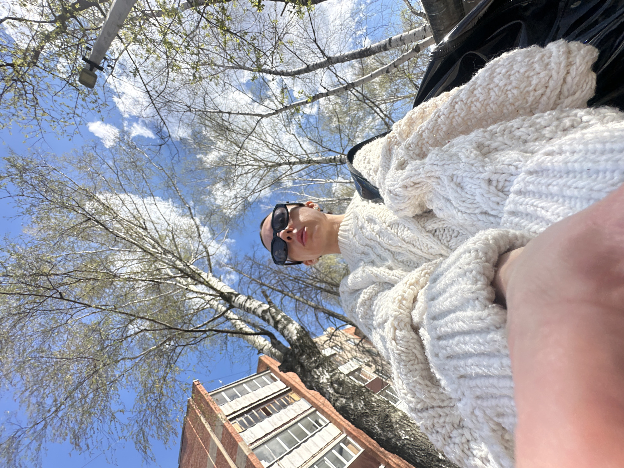 A low-angle shot looking up shows a person wearing a chunky white cable-knit sweater and sunglasses. The backdrop consists of a blue sky with scattered white clouds, the bare branches of trees with emerging green leaves, and the corner of a brick building with white-framed windows. A streetlight is visible in the upper left corner. The image appears to be taken in Votkinsk, Russia, as indicated by the architectural style and the type of trees common in the region. The person is looking slightly downward. The focus is primarily on the person and the immediate surrounding environment of trees and the building.