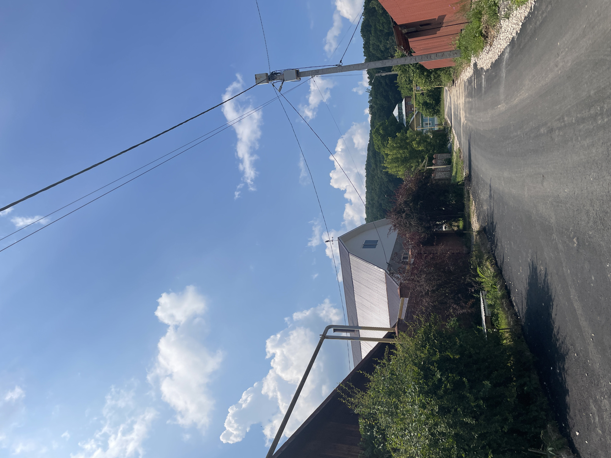 The image shows a residential street in a village. The sky is blue with scattered white clouds. A paved road runs through the center of the image, curving to the right. On the left side of the road, there is a house with a metal roof, a green bush, and a wooden structure. On the right side, there is a red corrugated metal fence and a utility pole with wires. In the background, there are more houses and a hill covered in trees. The scene appears to be a sunny day.