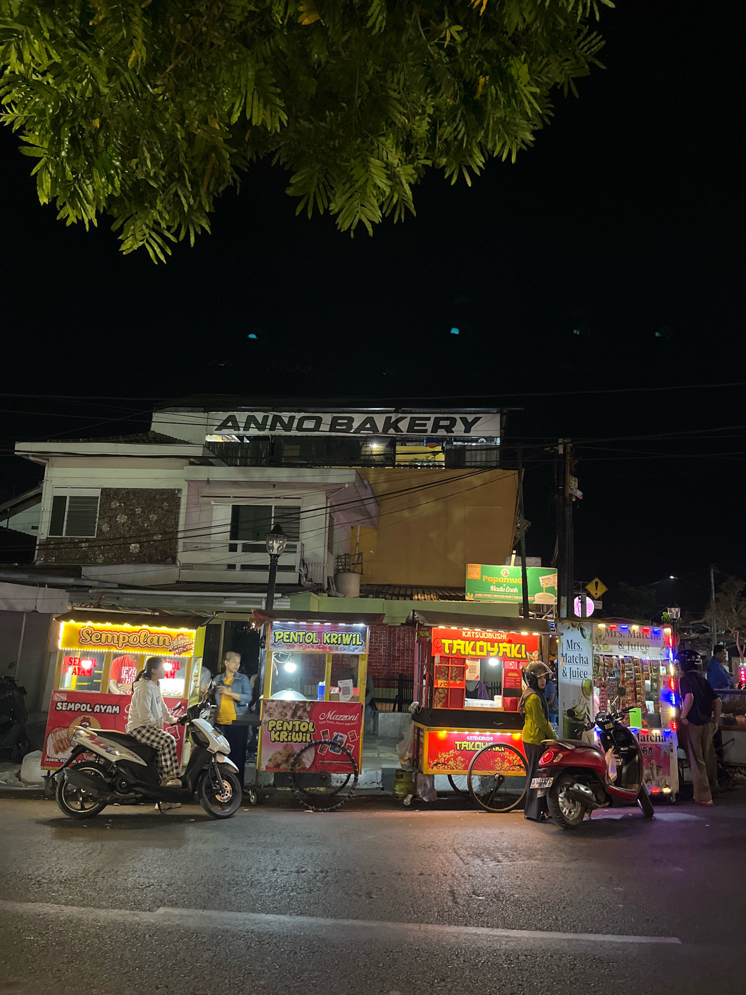 The image depicts a street scene at night in Salatiga, Indonesia. In the foreground, a paved road is visible with some white markings. Several food carts are set up along the roadside, illuminated by bright lights. The carts advertise various food items, including "Sempolan Ayam," "Pentol Kriwil," "Takoyaki," and "Mrs. Matcha & Juice."
A woman on a white scooter, wearing checkered pants and a light-colored hoodie, is stopped in front of the "Sempolan Ayam" cart. Two individuals appear to be customers or workers at the "Pentol Kriwil" cart. Another person, wearing a yellow top, is standing near a red scooter parked in front of the "Takoyaki" cart. Two men, one in a dark shirt and the other in a blue shirt, are visible near the "Mrs. Matcha & Juice" cart.
Above the food carts, a multi-story building is visible. A prominent sign on the building reads "ANNO BAKERY." The building has windows and some areas appear to be under renovation or have weathered facades. Green foliage from a tree hangs down from the top of the frame, obscuring a portion of the night sky. The overall atmosphere suggests a lively street food scene in the city at night.