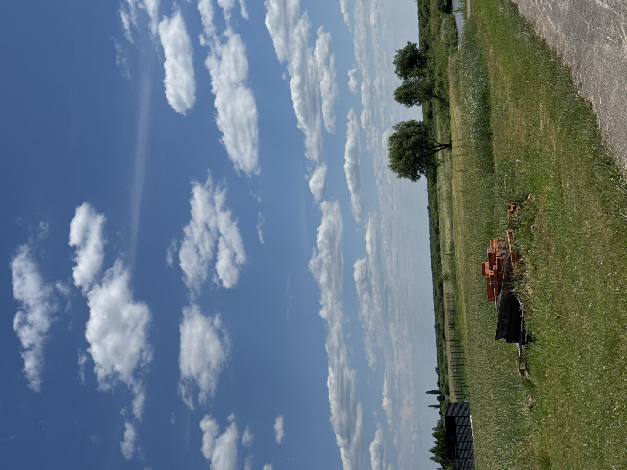 This is a vertical, outdoor photograph taken on a sunny day, likely in the late morning or early afternoon, given the bright light and clear blue sky dotted with fluffy white clouds. The scene depicts a rural landscape in Dovhe, Ukraine.
The foreground is dominated by a grassy field, with a paved path running along the right edge of the frame, suggesting a rural road or access point. Scattered within the field, towards the right side of the image, are some objects that appear to be building materials or debris, including what looks like a pile of reddish-orange bricks and a dark tarp-like material. A small, dark-colored shed or structure is visible towards the bottom left of the frame, with trees and vegetation extending into the distance.
Further into the middle ground, several trees are scattered across the landscape, their green foliage contrasting with the vibrant blue sky. A narrow body of water, possibly a canal or a small river, is visible in the far distance, lined with more trees and vegetation. A fence runs along the left side of the middle ground, delineating a boundary or an enclosure.
The sky is a prominent feature, filled with a variety of clouds, from small, wispy formations to larger, more defined cumulus clouds. The overall impression is one of tranquility and natural beauty, with a hint of human presence through the man-made elements. No people are visible in the image, and there is no visible text.