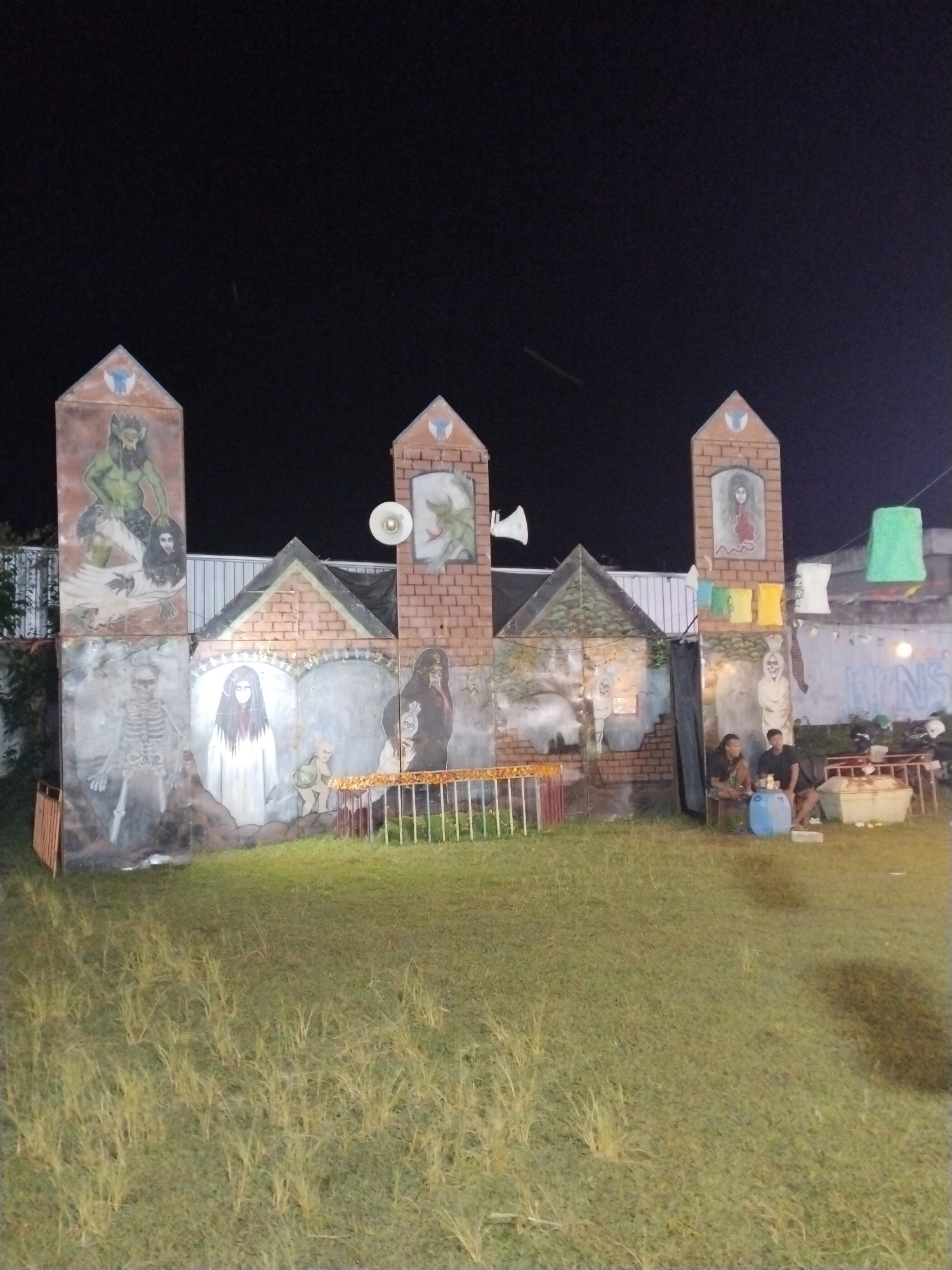 The image depicts an outdoor scene in Jetis, Indonesia, at night. The dominant feature is a makeshift castle-like structure with painted images of various ghoulish or monstrous figures, including skeletons, pale women with long dark hair, and other creatures. The structure is built of brick-patterned surfaces and adorned with triangular peaks. Attached to it are loudspeakers.
In front of the structure is a grassy area. To the right of the castle structure, two men are sitting, appearing to be resting. Near them are a blue barrel, a cooler, and a food vendor stand. The backdrop includes a concrete wall with graffiti and some hanging lanterns. The lighting is dim, with the structure and immediate area being lit.
