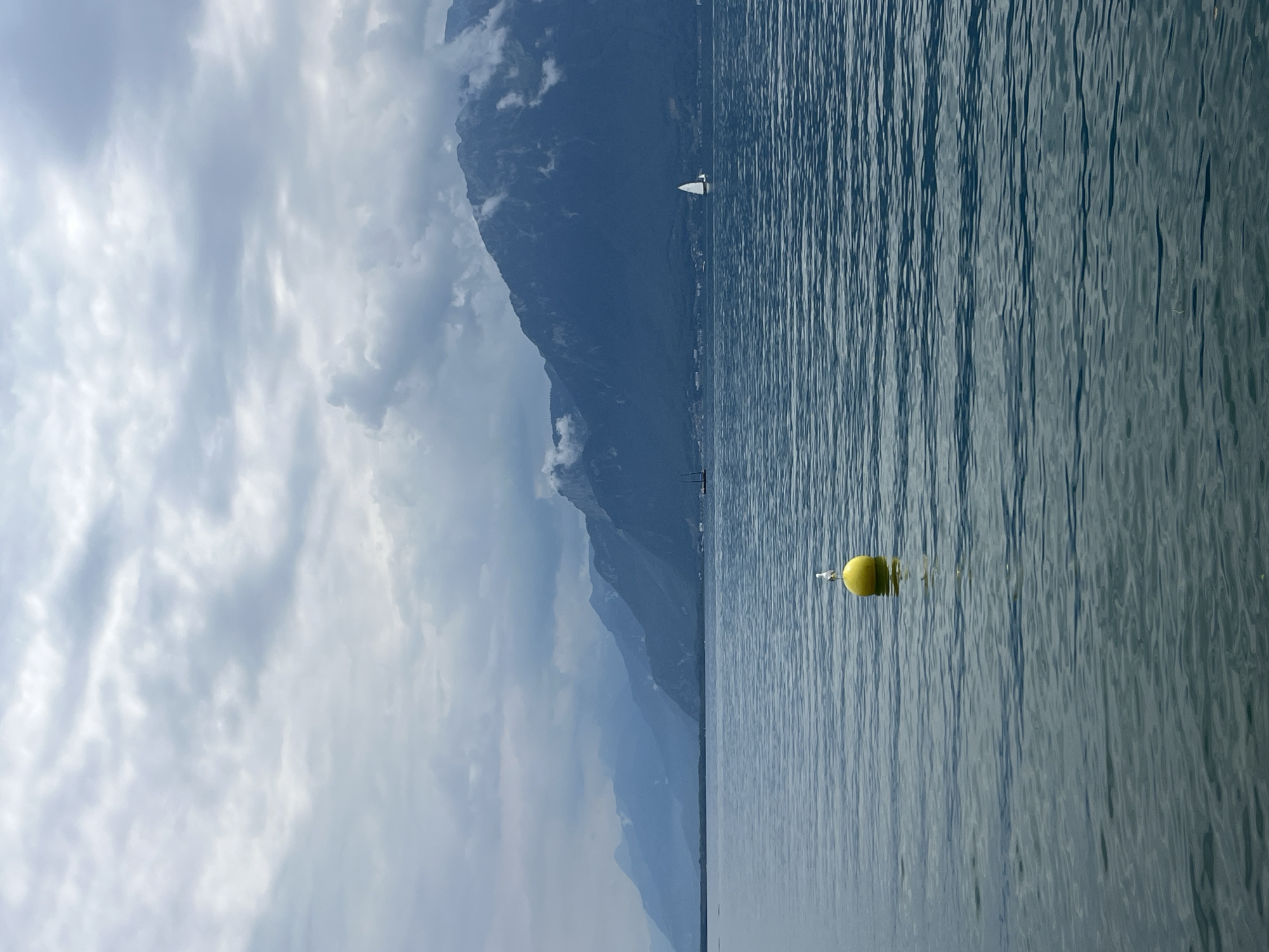 The image depicts a serene lake with a backdrop of majestic mountains under a cloudy sky. In the foreground, a bright yellow buoy floats on the calm water, with a single white bird perched atop it. The water has a rippling texture, reflecting the muted tones of the sky and mountains. Further out on the lake, two sailboats are visible, adding a touch of activity to the otherwise tranquil scene. The mountains in the distance are layered and shrouded in a soft haze, with some clouds clinging to their peaks. The overall atmosphere is one of peacefulness and natural beauty. This scene is likely Lake Geneva, with the Swiss Alps visible in the background. Vevey, Switzerland is a town situated on the northern shore of Lake Geneva, known for its stunning views of the lake and the surrounding mountains.