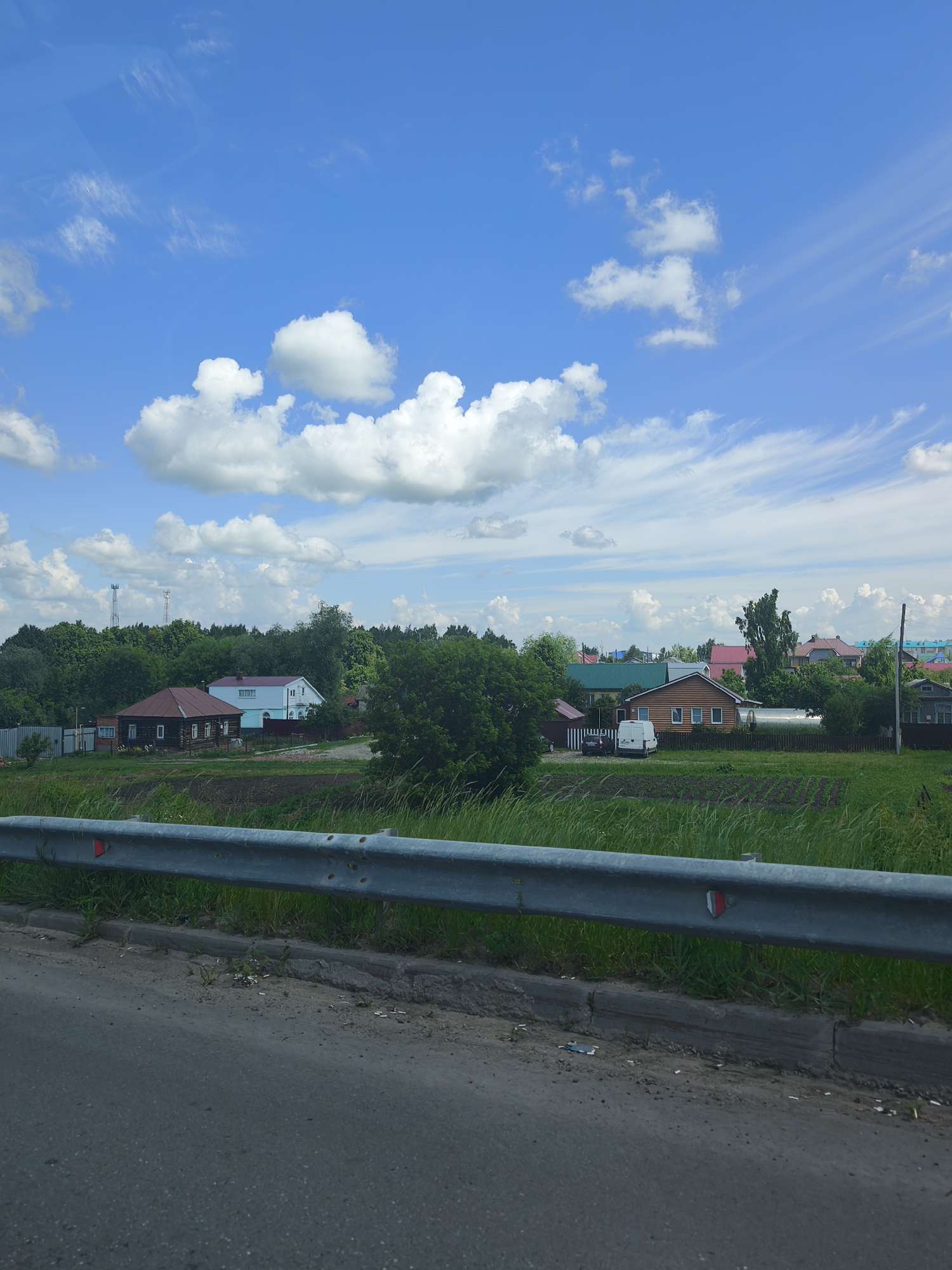 The image shows a rural landscape with houses and trees under a partly cloudy blue sky. A metal guardrail runs along the bottom of the frame in front of a grassy area and a field. Several houses, some with colorful roofs, are visible in the middle ground, interspersed with trees. There are also two radio towers in the distance. The foreground shows a paved road. The scene appears to be a typical residential area in a rural setting, possibly in the city of Kanash, Russia, given the style of the houses and vegetation. There are no people visible in the image.