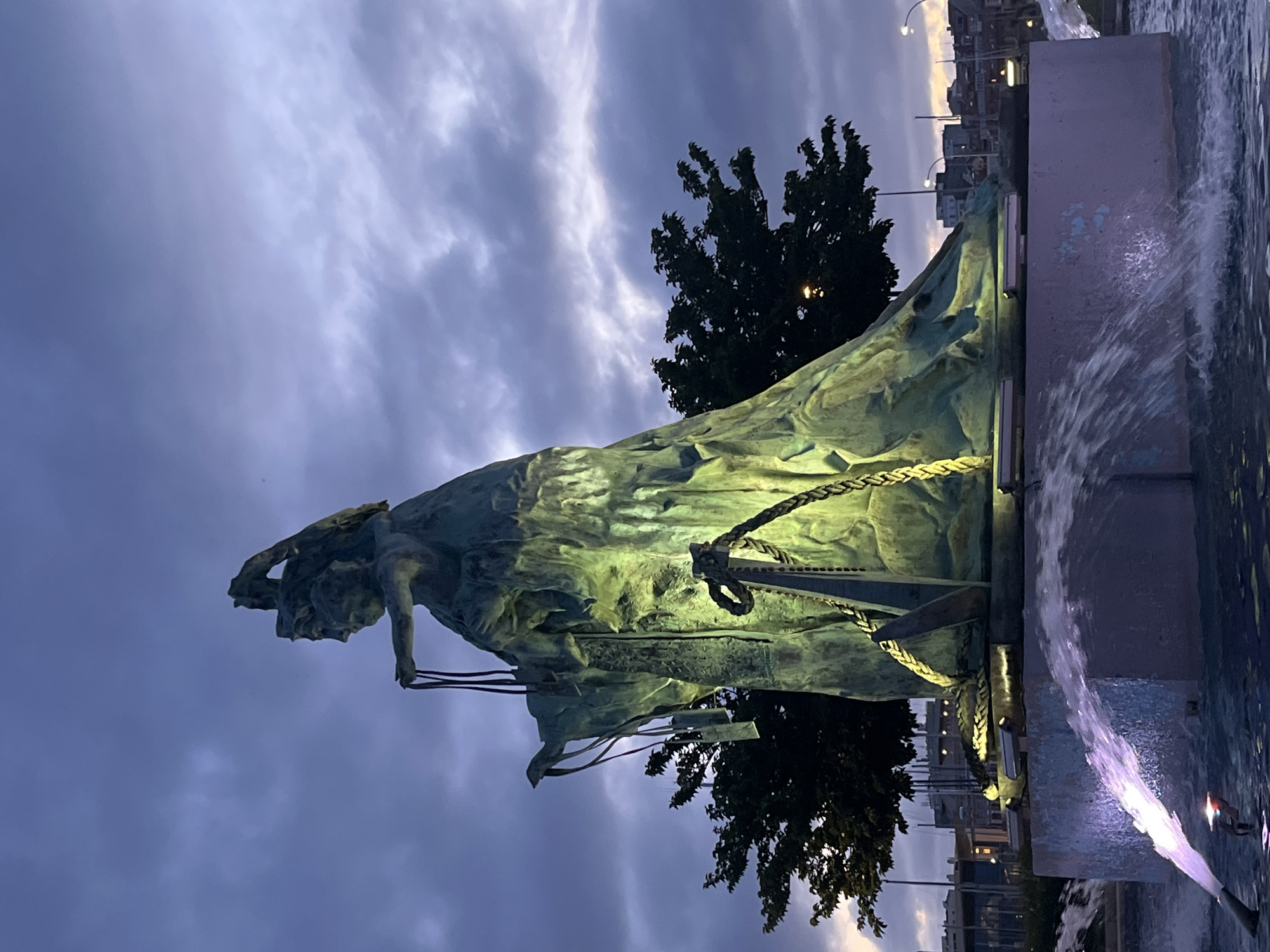 The image shows a nighttime view of the Emigrants' Monument in A Coruña, Spain. The sculpture is brightly illuminated, depicting a male figure straining at ropes, set against a backdrop of cloudy sky and a dark tree. Water cascades from the side of the monument's base, lit by embedded lights. In the background, buildings and streetlights are visible.