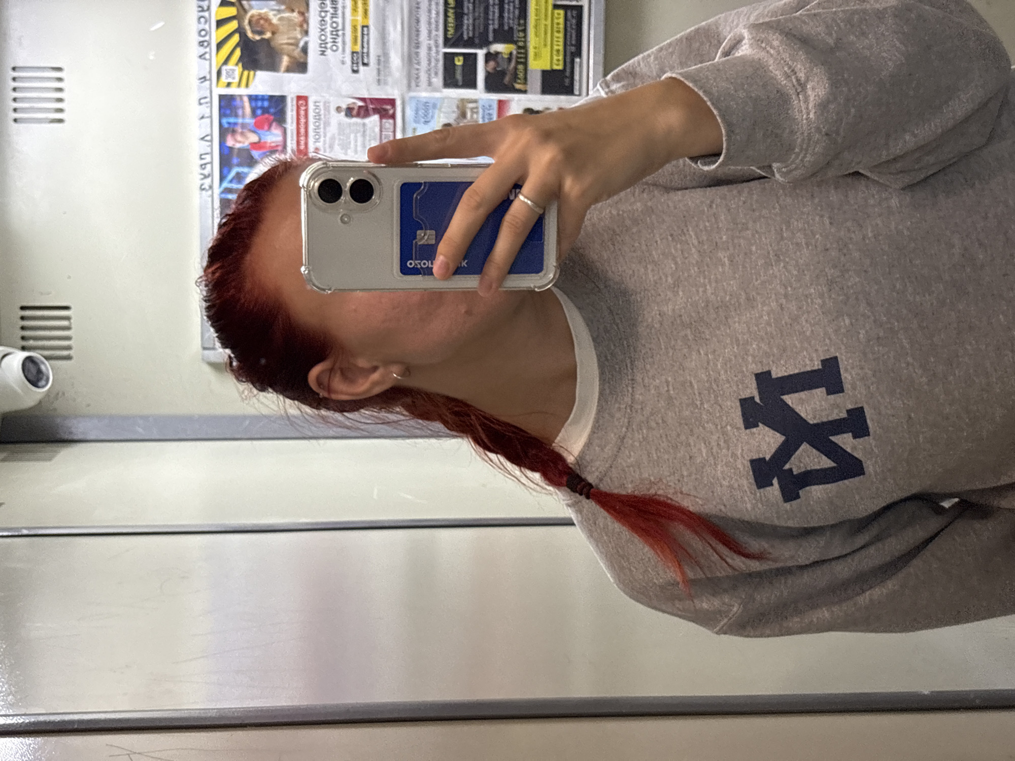 A young woman with red hair braided into a single plait is taking a selfie in an elevator. She is wearing a grey sweatshirt with a dark blue logo that appears to be an intertwined "M" and "Y". On the back of her phone, which is held up to take the photo, is a blue card holder with what appears to be a credit card inside. She is wearing a thin silver ring on her right hand. The elevator walls are a pale grey, and a security camera is visible at the top of the frame. In the background, reflected in the mirror of the elevator doors, is a bulletin board with various posters and notices, some of which appear to be advertisements. The posters are in Russian. The overall setting suggests an urban environment, possibly an apartment building or an office building in Rostov-on-Don, Russia.
