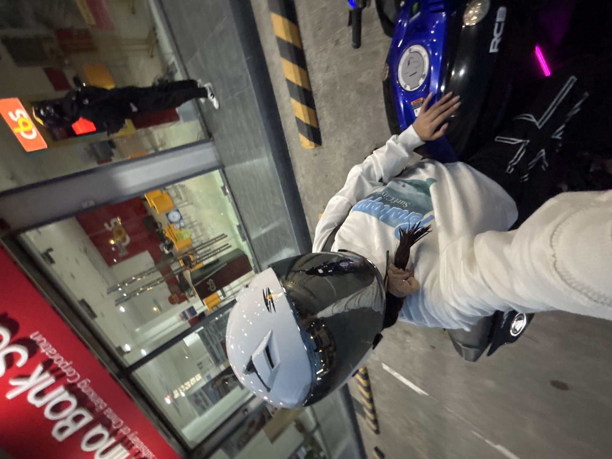 The image depicts a person on a motorcycle in front of a China Banking Corporation branch located in Davao City, Philippines. The rider is wearing a white helmet with a mirrored visor, a white long-sleeved shirt, and black pants with white designs. The motorcycle is blue and black, with "RCB" visible on the side. In the background, another person is standing outside the bank, wearing dark clothing and white sneakers. The bank's facade has a prominent red sign with white lettering. Yellow and black striped barriers are visible on the ground. The scene appears to be at dusk or night, with artificial lighting illuminating the area.