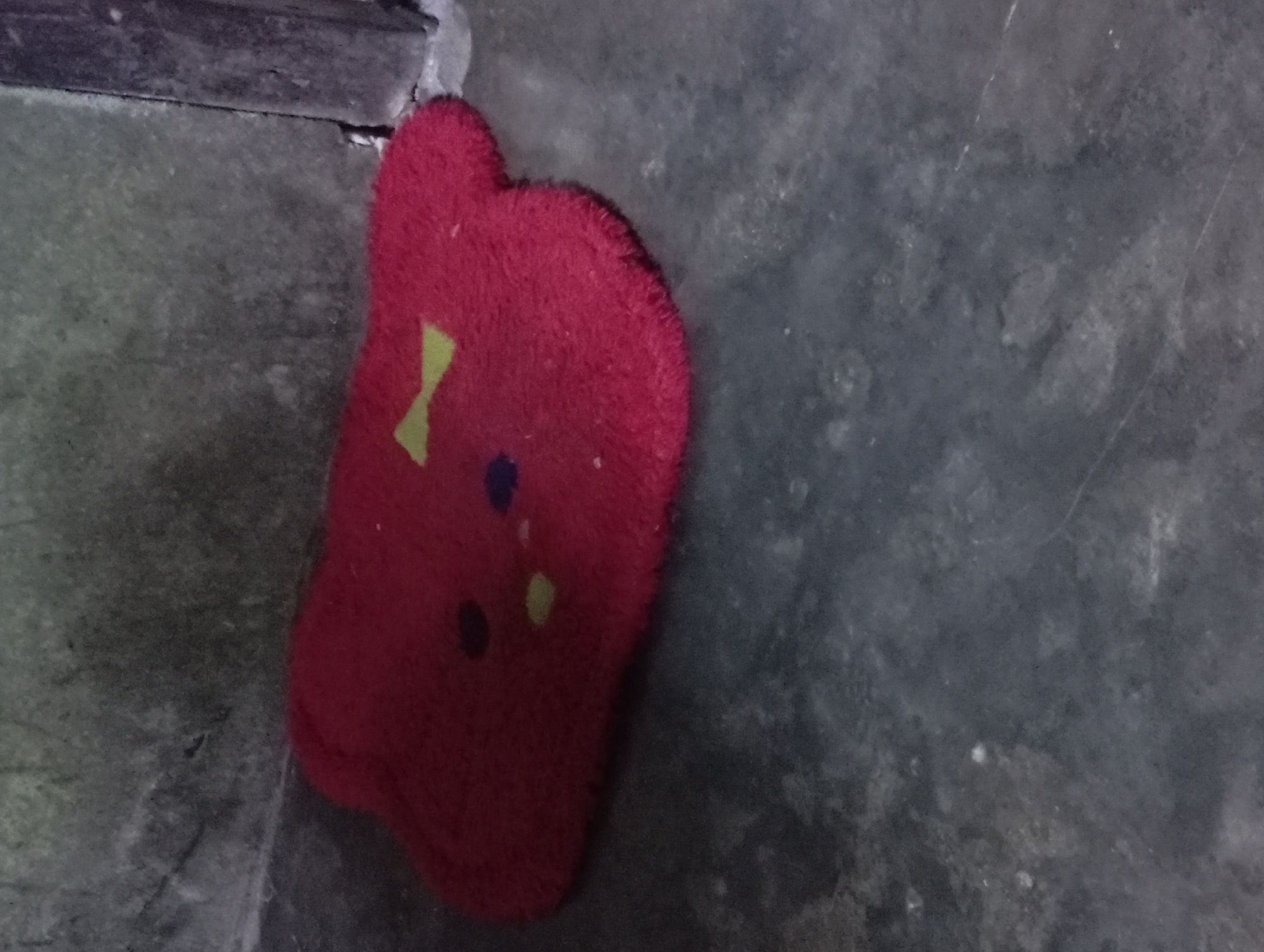 A close-up, slightly blurry, overhead shot shows a red, fuzzy mat on a dark, uneven concrete floor. The mat is shaped like a simple cartoon bear's head, with two rounded ear-like protrusions at the top. A yellow, bow-tie shape is centered on the "forehead" area of the mat, and two small, round, dark blue spots are positioned below it, resembling eyes. The floor appears to be dirty and worn. A dark vertical surface, possibly a doorframe or wall, is visible on the far right edge of the image, slightly out of focus. The scene is dimly lit, casting subtle shadows. The context provided places this scene in the city of Rejosari, Indonesia.