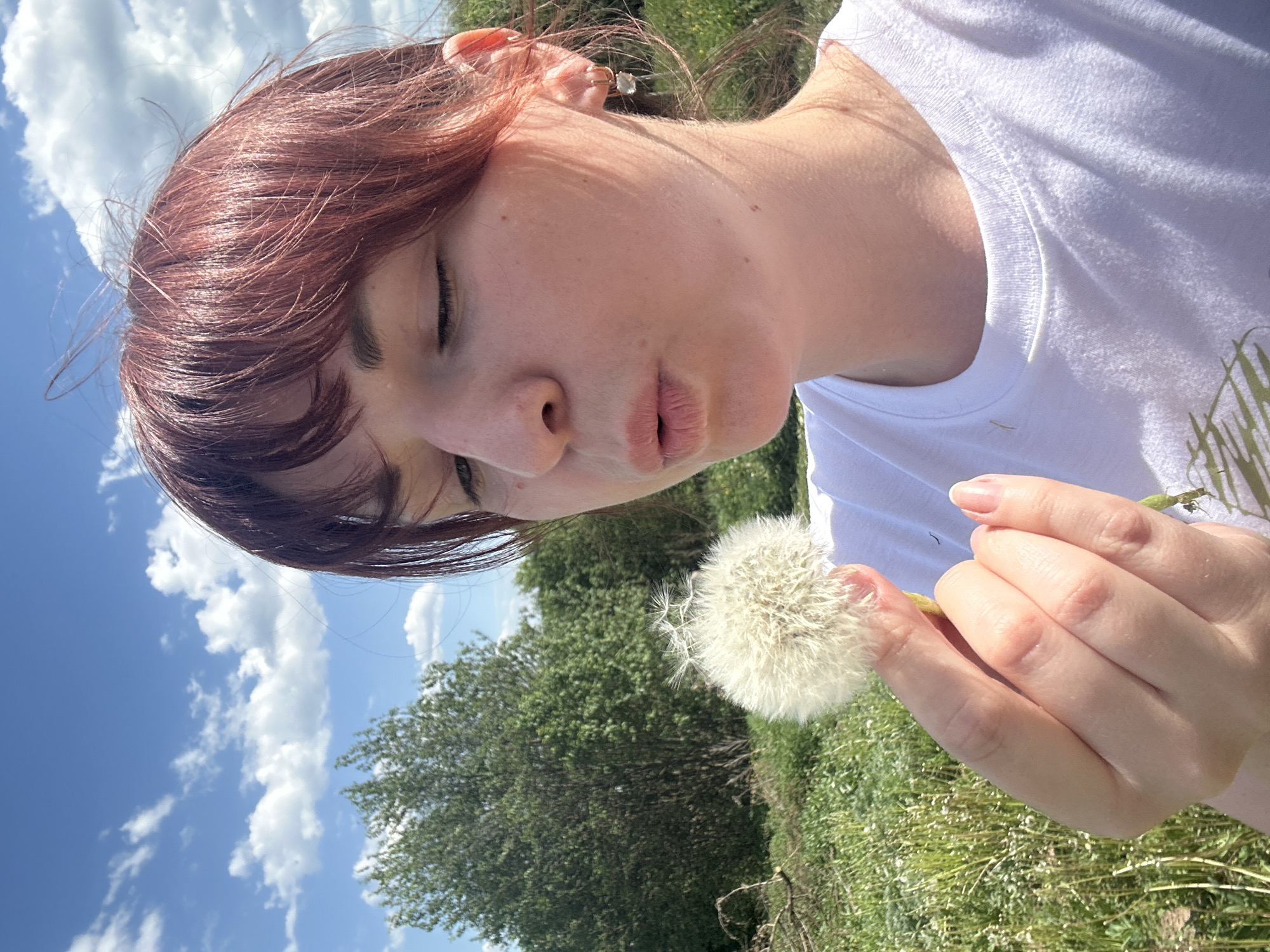 A woman with reddish-brown hair stands outdoors in a field, potentially in or near the city of Фоки, Russia. She holds a dandelion puff in her right hand and appears to be blowing on it, with her eyes closed and lips puckered. She is wearing a white t-shirt. The background consists of green trees and grass, a blue sky, and white clouds. The image is shot from a low angle, looking up at the woman.