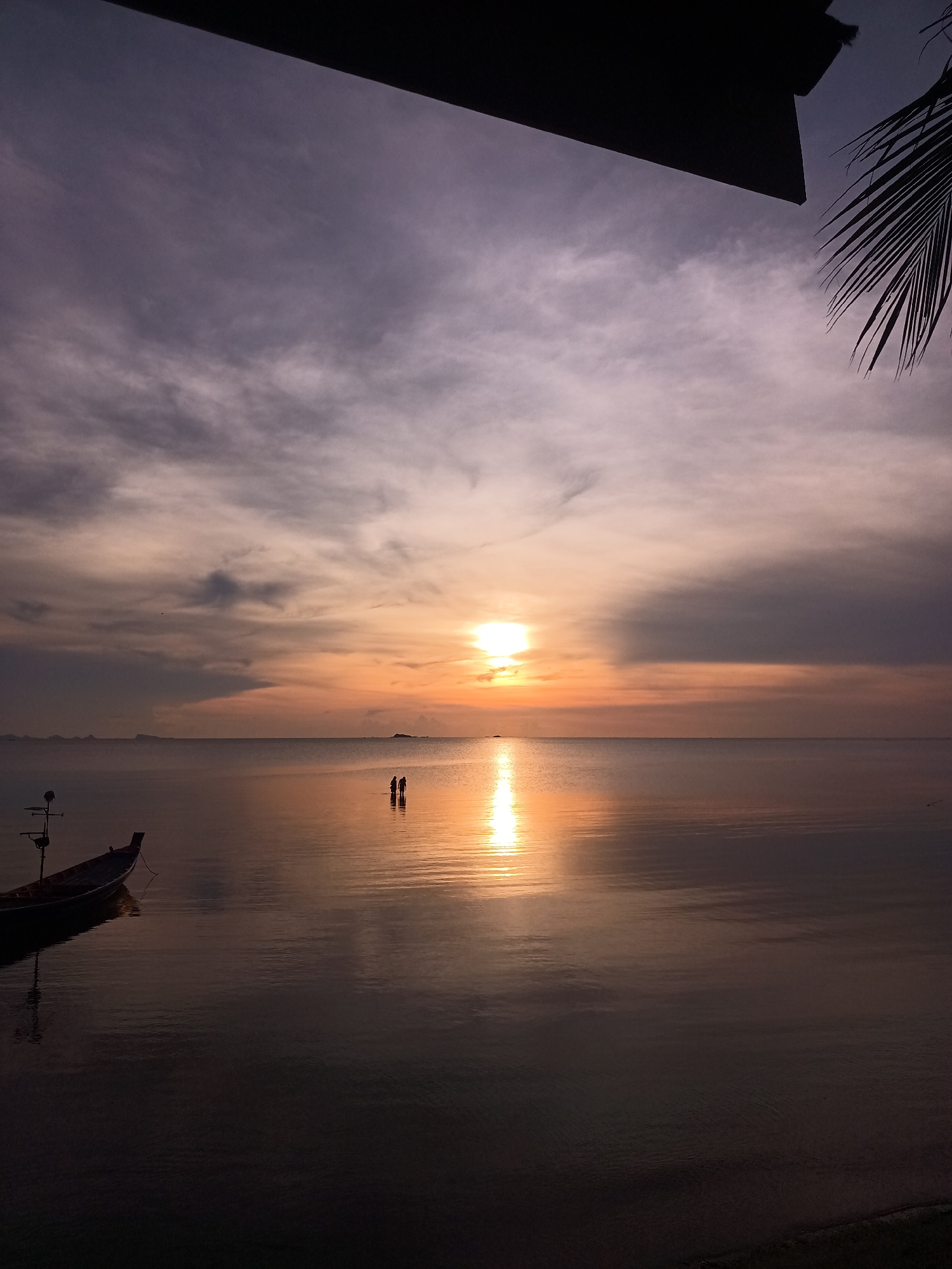 The image captures a sunset at Baan Wok Tum, Thailand. The sky is filled with clouds colored in hues of purple, orange, and gray by the setting sun. The sun's reflection creates a bright, orange path across the calm water surface. In the distance, two silhouetted figures stand in the water. A small fishing boat is moored to the left of the frame. The bottom edge of a structure with a palm frond is visible at the top right of the image, and a dark roof line cuts across the top.