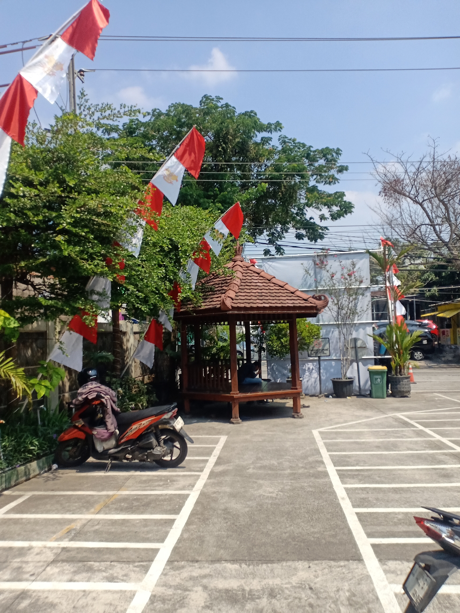 The image shows a parking lot scene in Semarang, Indonesia. The parking lot is paved and marked with white lines. In the foreground, a red and black motorcycle is parked. In the background, there is a small gazebo with a brown tiled roof. Red and white flags are strung across trees and poles. A person is sitting inside the gazebo. There are also several potted plants and trash cans. A building with a white tarp covering it is visible behind the gazebo. The sky is clear and blue. The overall impression is that of an outdoor area, possibly a public space or the grounds of a business, decorated for a celebration or event.