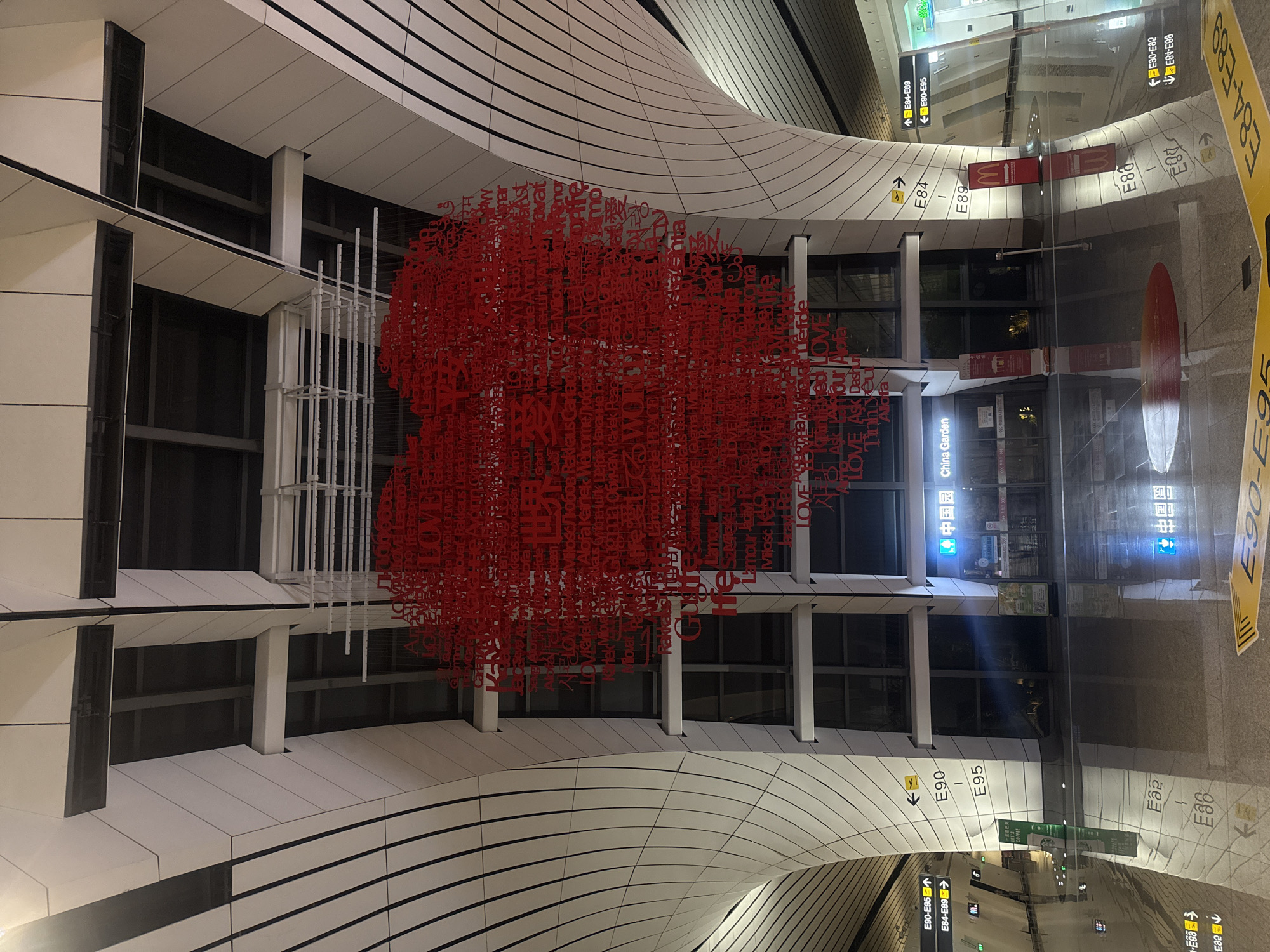 An interior shot of a modern airport terminal features a large, red heart-shaped art installation suspended from the ceiling. The heart is composed of numerous words written in various languages, including "LOVE," "Peace," and "Amour." The installation is illuminated by ambient lighting.
The terminal itself has a sleek, futuristic design with curved white walls and a high ceiling. Below the art installation, there are large windows that reveal a darker interior space. Signage is visible on the walls, including directional arrows and gate numbers like "E90-E95." A McDonald's sign is also present.
The floor is highly reflective, mirroring the architecture and the art installation. On the floor, yellow markings and arrows indicate pathways, with "E90-E95" being clearly visible. A sign with "China Garden" is displayed on one of the storefronts. The overall environment suggests a busy public transportation hub. The location is Guangyang District, China.