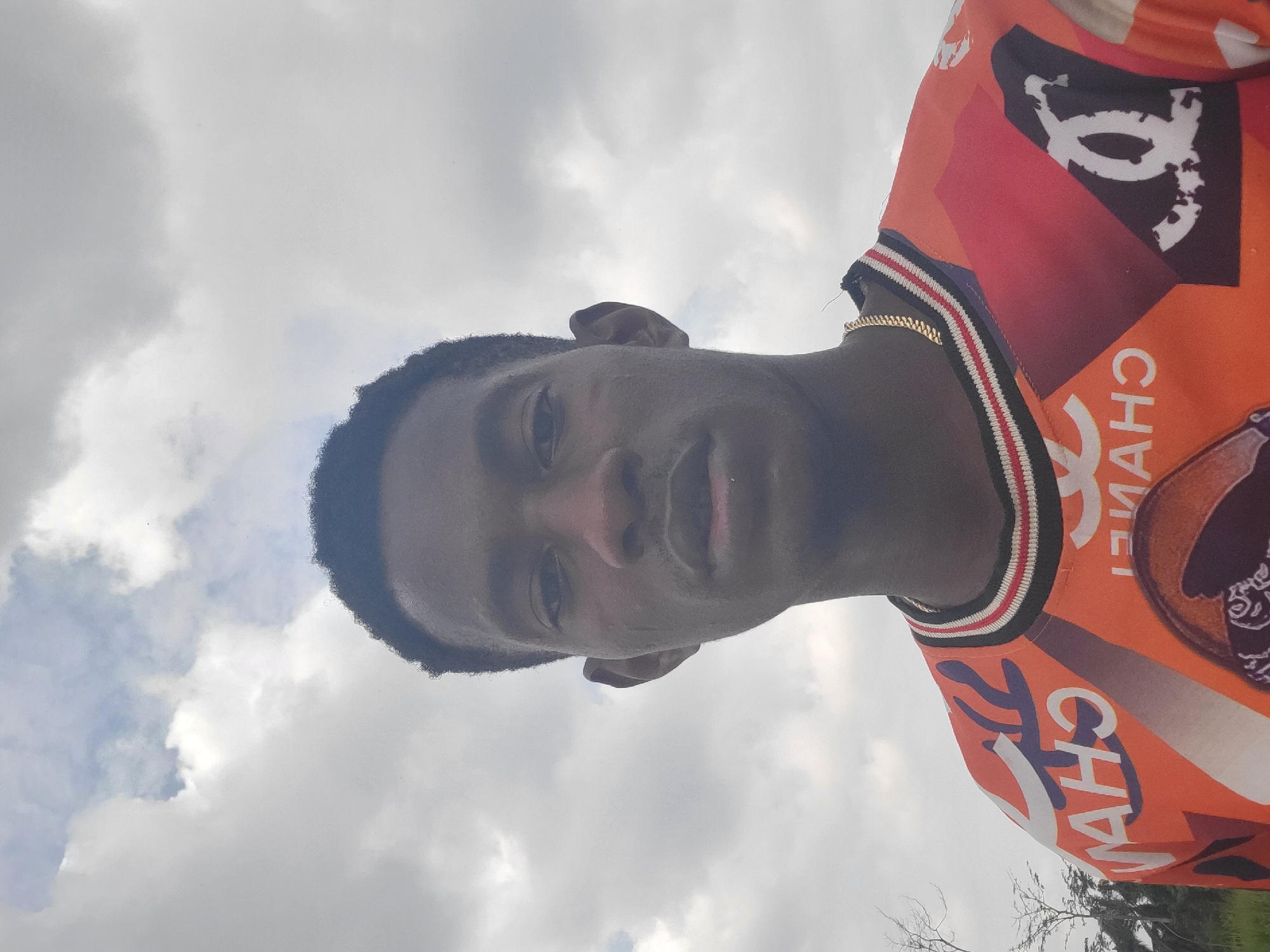 The media file shows a young Black male against a cloudy sky. He is wearing an orange t-shirt with a repeating "CHANEL" logo pattern in white and black. The collar of the t-shirt has a striped band with red, white, and black stripes. He is also wearing a gold chain. The image is a close-up, with the subject's face filling a significant portion of the frame. The lighting suggests it is daytime, and the cloudy sky indicates overcast weather. Some tree branches are visible in the lower right corner of the image. The overall mood is neutral, with the subject looking directly at the camera. The image appears to be taken outdoors in Egbagu, Nigeria.