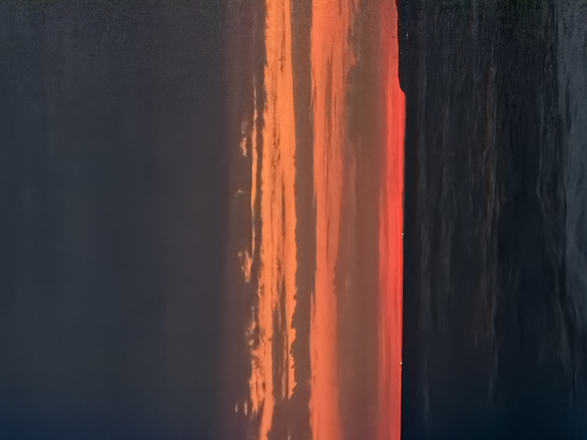 The image depicts a dramatic sunset over the Black Sea, with the coast of Sochi, Russia visible in the distance. The sky is a gradient of dark gray at the top, transitioning to vibrant orange and red hues where the sun is setting. Wispy clouds are scattered across the orange band, catching the last rays of light. Below the horizon, the sea is dark and choppy, reflecting the dim light of the sky. A faint line of lights from the distant shore of Sochi can be observed. The overall scene is serene and evokes a sense of calm before nightfall.
