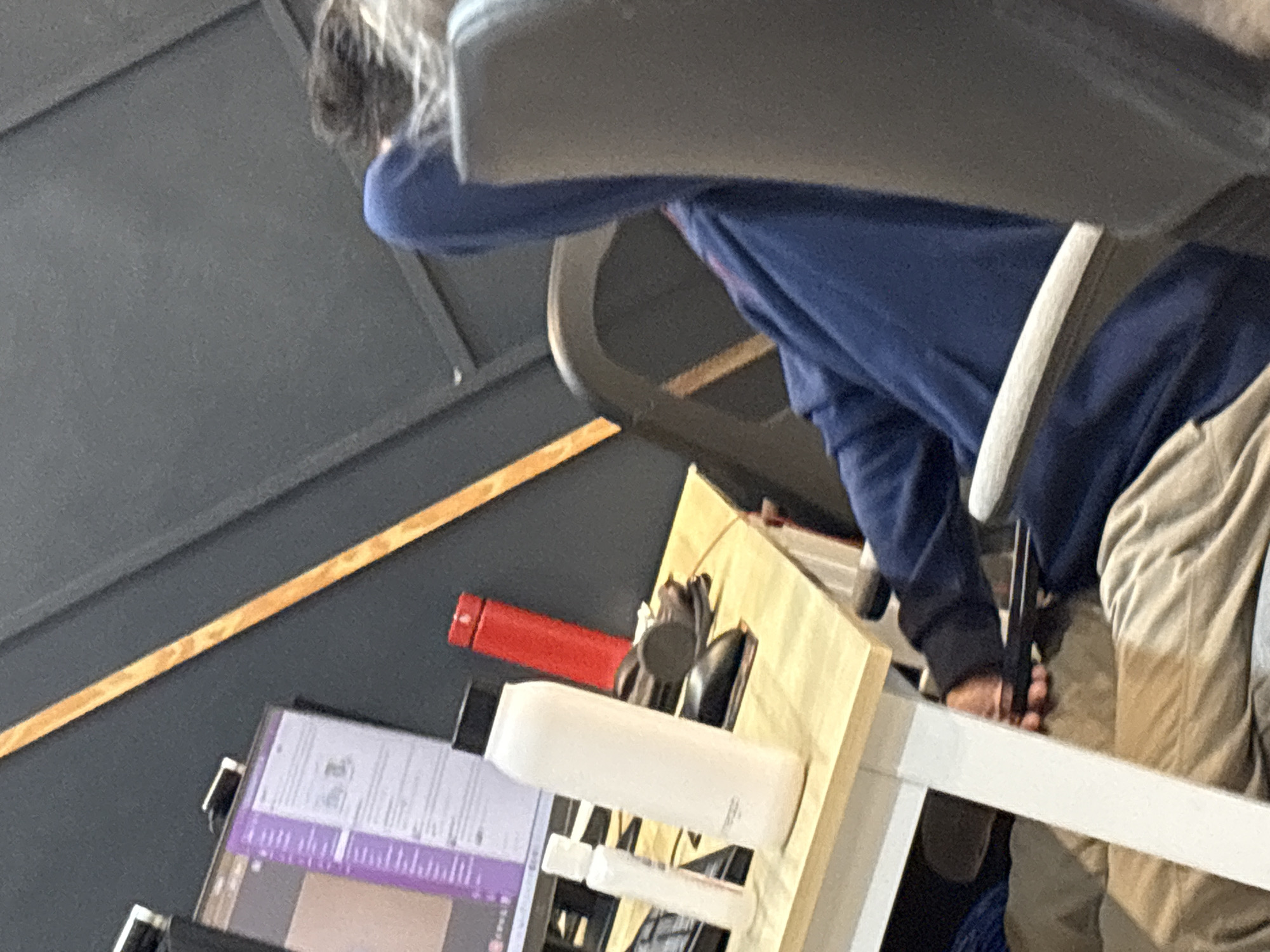 A person is seated at a desk in an office environment. The person is wearing a blue hooded sweatshirt and beige pants. Their head is tilted down, obscuring their face, and their hands are not visible. On the desk, there is a white water bottle, a red thermos, and a computer monitor displaying a purple interface. The office has dark gray walls with a diagonal wooden beam. The image is a close-up, with the person and desk dominating the frame. The location is described as Davao City, Philippines, though there are no specific visual cues to confirm this.