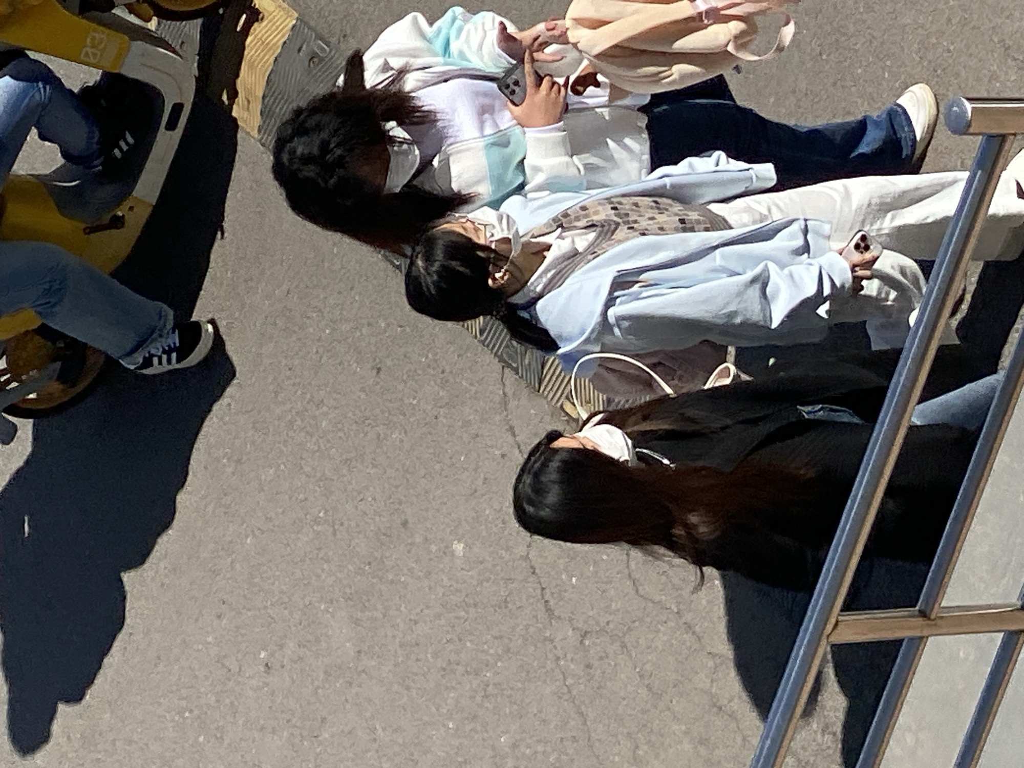 The image, captured from an elevated perspective, shows three young women standing on a gray concrete surface. All three women are wearing face masks. One woman, wearing a white and light blue hoodie, is holding a smartphone. Another woman is wearing a light blue jacket over a patterned top and white pants, also holding a smartphone. The third woman has long, dark hair and is wearing a dark jacket. To the left, a person's legs are visible on a yellow and white scooter. To the right, a metal structure is present. The location of Zhongtun, China is indicated by the prompt but cannot be independently verified from the image alone.