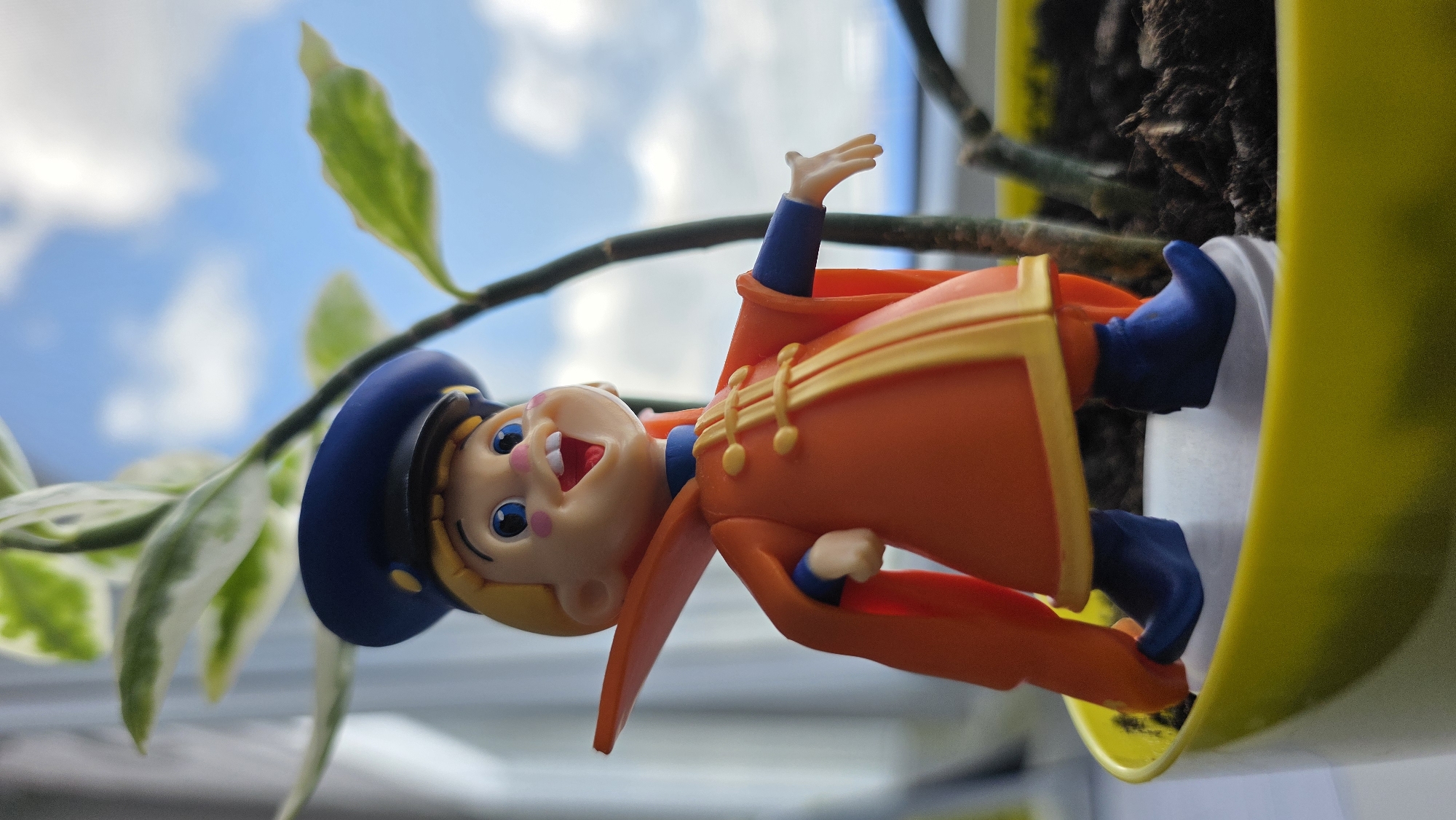 A small, plastic figurine of a cartoon character stands in a yellow pot with a plant. The figurine is dressed in an orange coat with yellow trim and a blue hat. It has large blue eyes, a wide smile, and pink cheeks. The plant has green and white variegated leaves. The background shows a blurred view of a blue sky with white clouds. The scene appears to be indoors, possibly near a window. There is no indication of the location in the city of Brest, Belarus.
