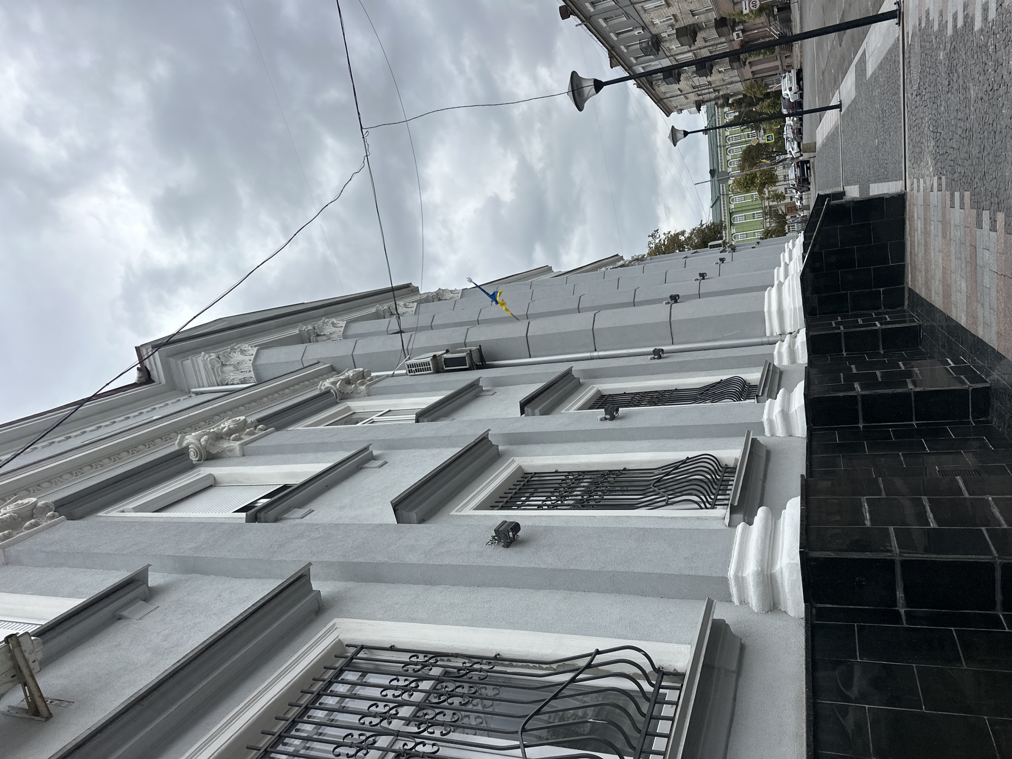 The image shows the facade of a building in Ternopil, Ukraine, on an overcast day. The building is light gray with white decorative columns and window frames. The windows have black wrought-iron bars. A Ukrainian flag is visible on the side of the building. The street in front of the building is paved with cobblestones. Several cars are parked on the street, and streetlights line the sidewalk. In the background, another building with a green facade and arched windows can be seen. The overall scene is urban and architectural, with a somber atmosphere due to the cloudy sky.