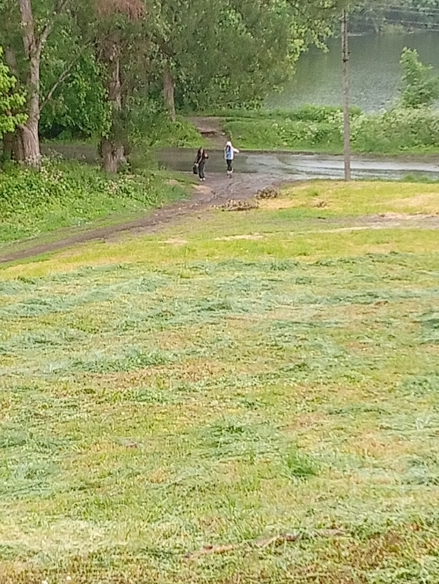 The image shows a grassy field in Poltava, Ukraine. In the background, two people are walking across a wet road or path near a body of water. One person is wearing a white jacket and dark pants and the other person is wearing dark clothing. The grass in the foreground appears freshly cut. Trees line the background beyond the road. A utility pole is visible near the water's edge.
