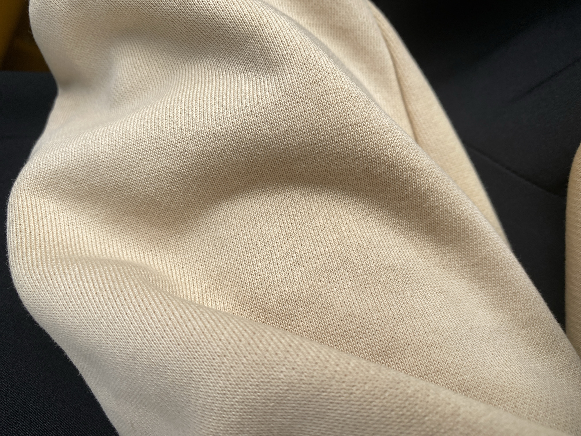 The media file is a close-up, still image showcasing the texture of a light beige, knitted fabric, likely a sweater or sweatshirt. The fabric is draped and folded, creating soft curves and shadows that highlight its ribbed pattern. The texture is fine and dense, suggesting a comfortable and warm material. In the background, a dark fabric, possibly black, provides a stark contrast to the light beige, further emphasizing the texture of the main subject. The lighting appears to be soft, creating gentle gradients across the fabric's surface. There are no people, animals, or distinct objects visible, focusing entirely on the textile's tactile qualities. The image does not convey any specific activity or event, but rather a still life composition centered on the material. No text is visible in the image. The context provided, Jalingo, Nigeria, offers a geographical location but does not directly influence the visual elements presented in this close-up view of the fabric.