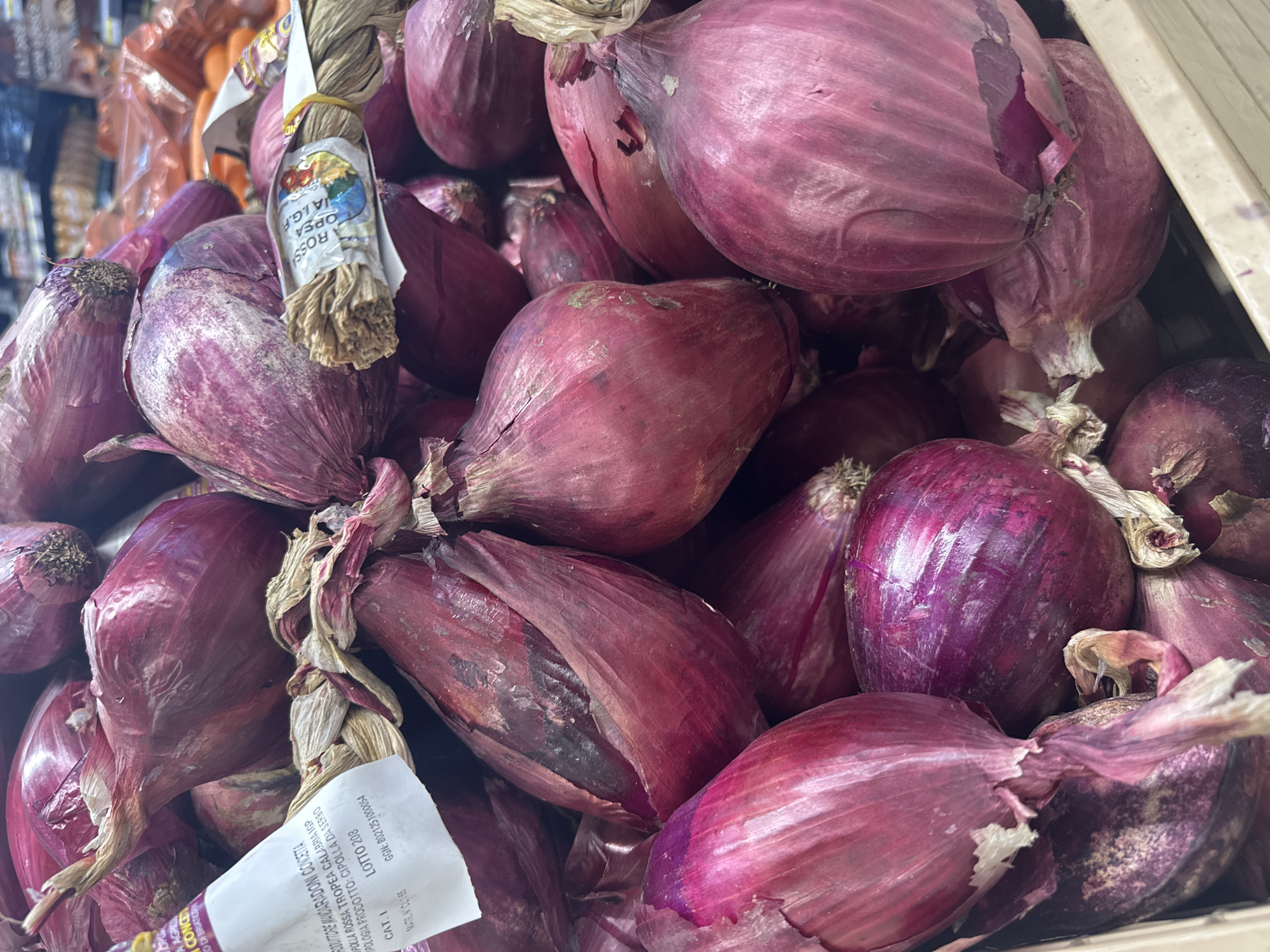 A close-up, high-angle, full-frame shot shows a pile of red onions in a grocery store in Trieste, Italy. The onions are mostly a deep red-purple color with some lighter pink and white layers visible. Some onions have dried brown stems and papery outer skins that are peeling away. A white label with Italian text and a barcode is attached to one bunch of onions. Another band around a bunch of onions has an image of a farmer and the text "CIPPOLLA ROSSA TROPEA CALABRIA IGP". The onions are densely packed in a light brown plastic bin. In the blurred background, other produce and shopping aisles are visible.