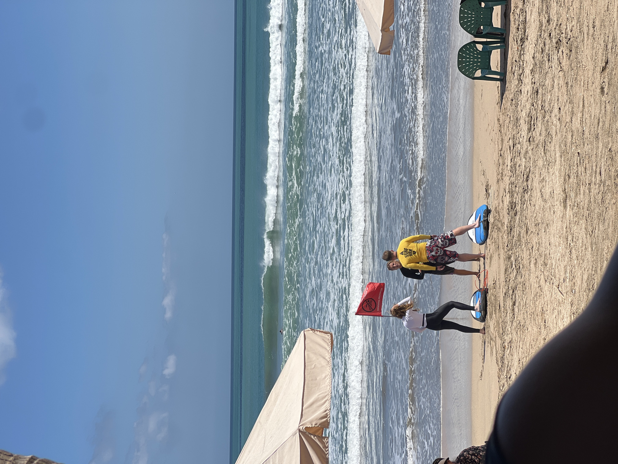 This image depicts a bright, sunny day at a beach in Kuta, Indonesia. The scene is set with the ocean in the background, featuring moderate waves and a clear blue sky. In the mid-ground, three individuals are standing on the sandy beach, appearing to be in a surfing lesson or activity. Two people are facing the camera, while one person is facing away. One of the individuals, a woman with long, blonde hair, is wearing a white long-sleeved shirt and black leggings, standing on a blue and white surfboard. Beside her, a man in a yellow rash guard with "SURF VAN" printed on it and patterned shorts stands on another surfboard. Between them, a third person in a black rash guard stands. A red flag with "SWIMMING PROHIBITED" written on it is planted in the sand nearby, indicating potentially dangerous conditions. Beach umbrellas, a common sight in Kuta, are visible on the left and right sides of the frame, providing shade. Two green plastic chairs are also positioned on the sand, suggesting a relaxed atmosphere. The foreground shows a person's arm and leg, out of focus, implying the photographer is also on the beach. The overall impression is of a typical day at a popular surf destination.
