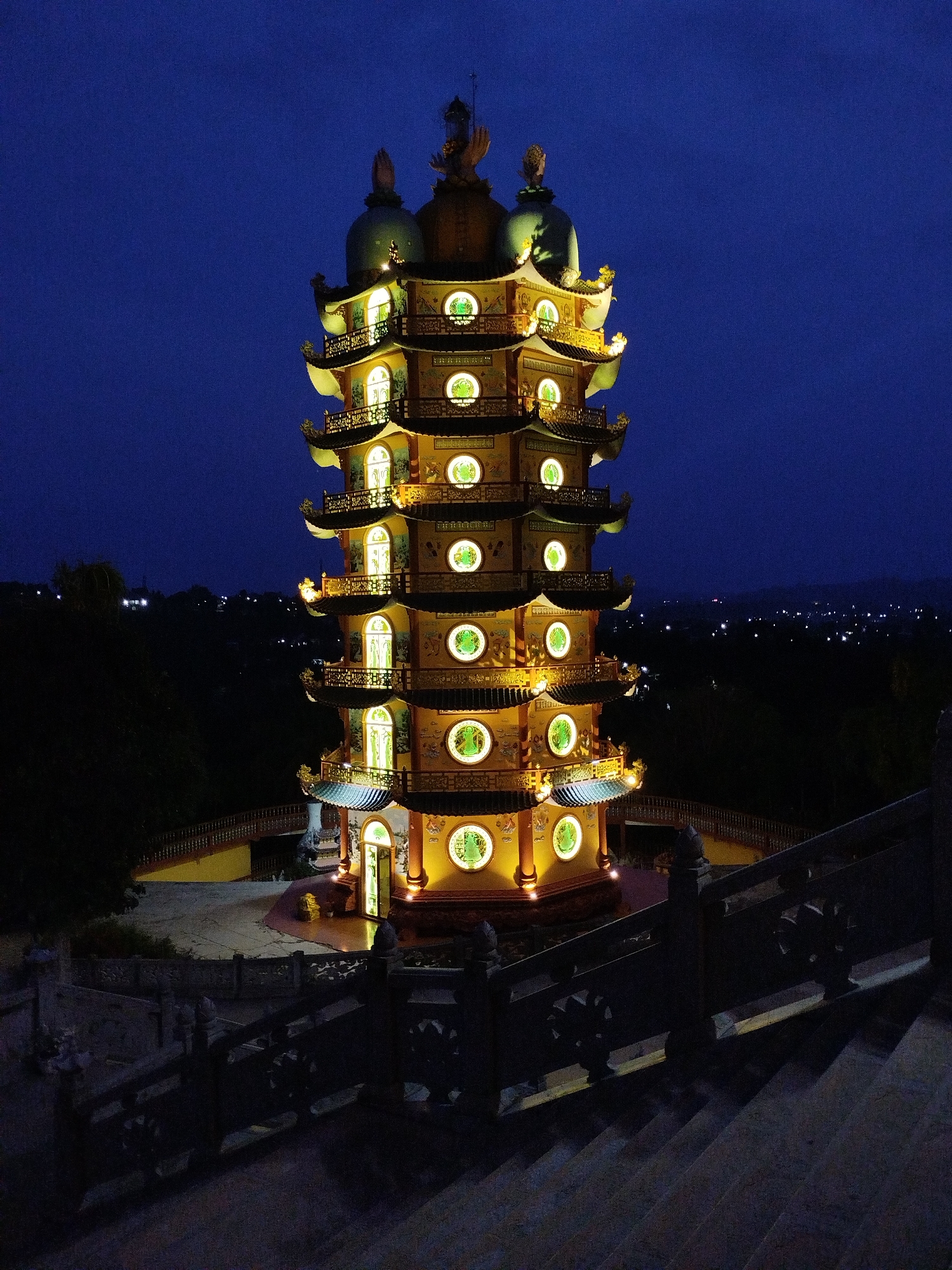 The image depicts the illuminated Bửu Long Temple in Phú An, Vietnam, at night. The multi-tiered pagoda is centrally positioned, its golden facade contrasting against the dark blue sky. Each level features circular windows emitting a soft, warm glow, and traditional architectural details like curved eaves and decorative embellishments. The pagoda sits atop a paved platform enclosed by railings. A flight of stairs with intricately patterned stone railings leads towards the structure. The background reveals a dark landscape with scattered city lights in the distance, suggesting a vantage point offering a view of the surrounding area. The foreground is defined by the descending staircase, subtly indicating the viewer's perspective from a higher elevation.