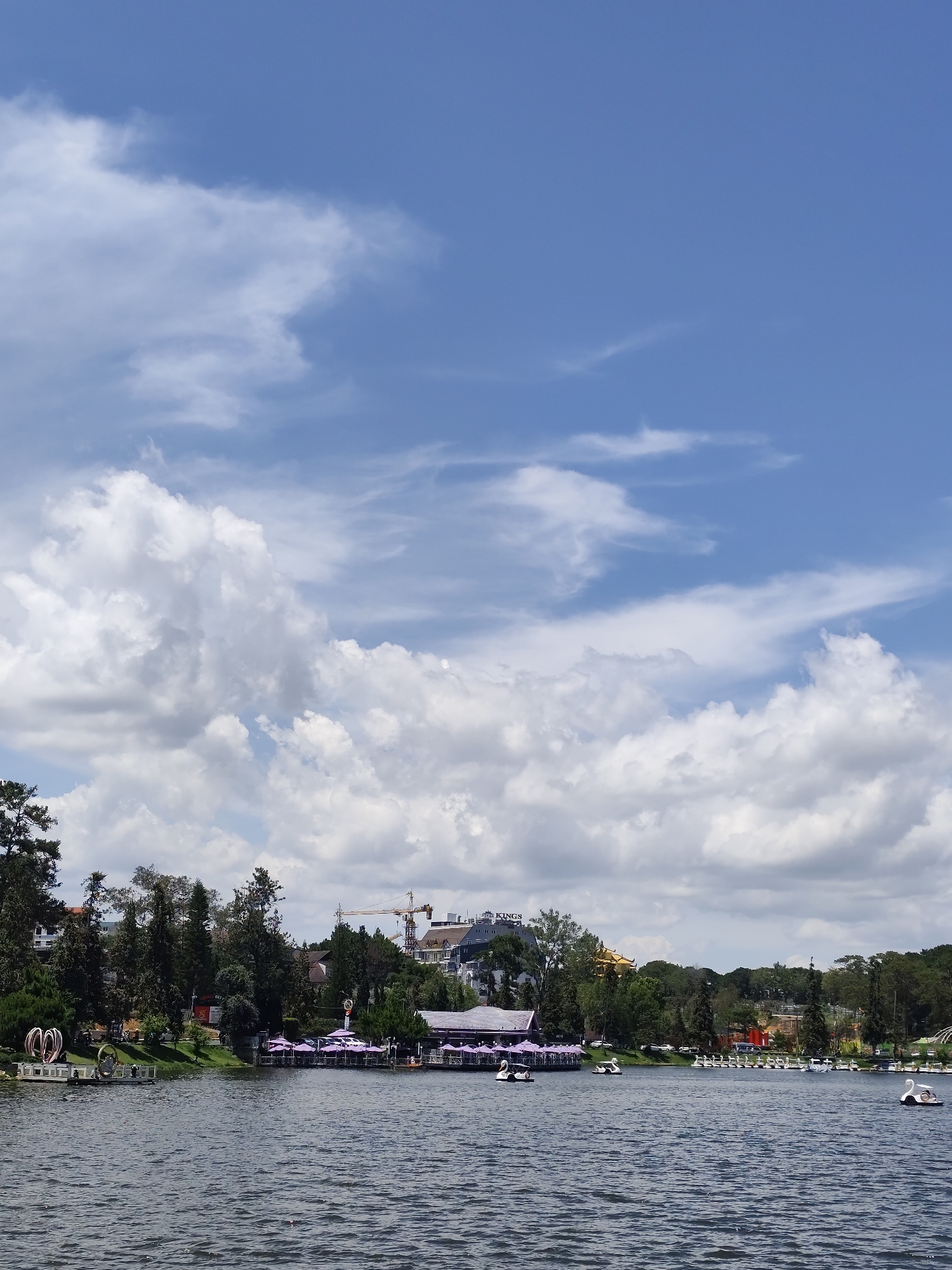 This image depicts a scenic view of a large lake, likely Xuan Huong Lake in Da Lat, Vietnam, under a bright blue sky with scattered white cumulus clouds. The scene conveys a pleasant, sunny day, likely during late morning or early afternoon.
The foreground is dominated by the dark, rippling surface of the lake. Along the distant shoreline, a verdant band of trees, including tall conifers, frames various structures. On the left side, near the water, abstract sculptural elements resembling intertwined hearts or rings are visible. Adjacent to these, a structure resembling a restaurant or cafe features numerous uniform purple patio umbrellas extending along the lake's edge, suggesting an outdoor seating area.
Further along the shore, a multi-story building with the text "KINGS" clearly visible on its facade stands prominently. A tall yellow construction crane is positioned next to this building, indicating ongoing development or renovation in the area. Other buildings and houses are interspersed among the trees.
The primary activity taking place on the lake is recreational boating. Numerous pedal boats, including several distinct swan-shaped ones, are scattered across the water. Some boats are actively being used by individuals or small groups, while others appear to be docked or waiting. The overall atmosphere is one of leisure and enjoyment at a popular destination.