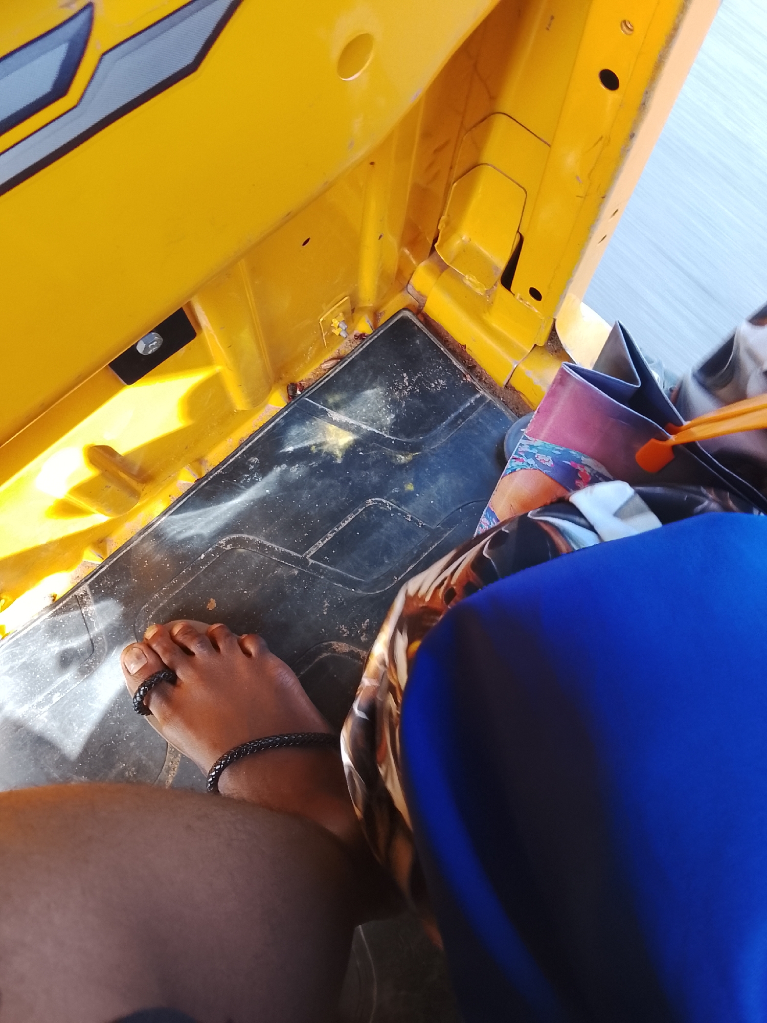 The image shows a close-up view of the interior of a yellow vehicle, possibly a tuk-tuk or auto-rickshaw, with a person's right foot visible on the floorboard. The floorboard is dark and appears worn, with some dust or debris. The person's foot is bare, with a dark braided ring on the big toe and a matching braided bracelet on the ankle.
The interior of the vehicle is bright yellow and shows some of its structural components, including bolts and seams. Sunlight is casting strong shadows on the yellow surfaces, indicating it's daytime. In the background, partially visible, are bags and what appears to be a person's legs covered in blue fabric, suggesting there might be another occupant or personal belongings. The overall impression is that of being inside a vehicle in motion, as suggested by the slightly blurred background. The context provided, "Tabari Jauro Manu, Nigeria," places the scene geographically. No text is visible in the image.