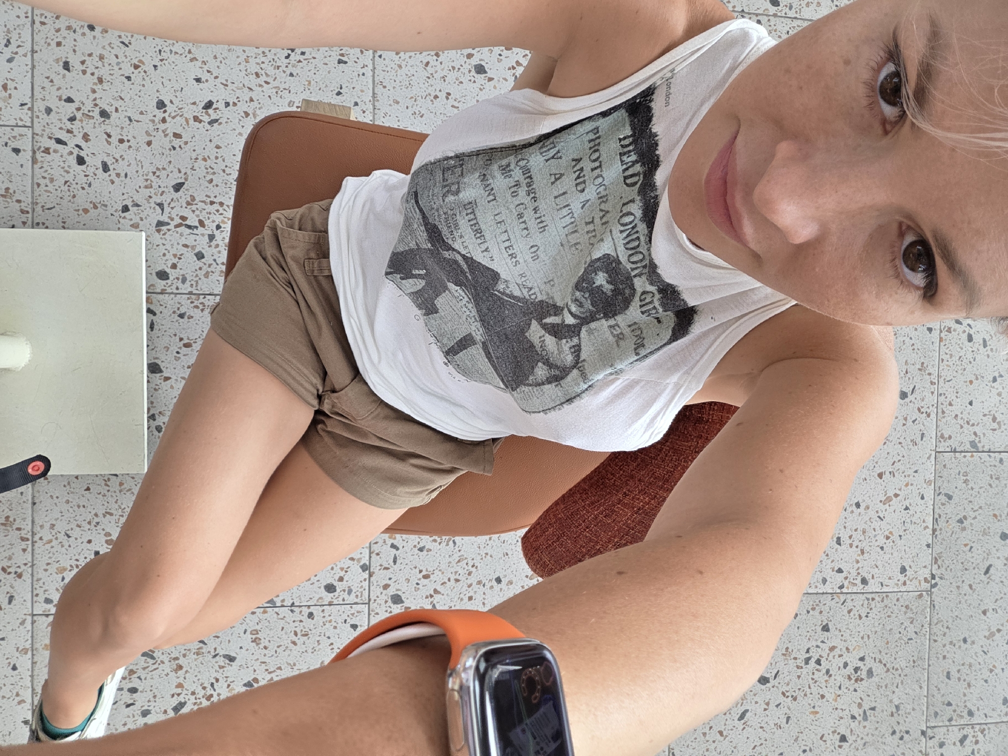 A woman wearing a white tank top with a graphic print, khaki shorts, and a smartwatch sits on a brown chair. The floor is tiled with a speckled pattern. A white table with a cylinder on it is visible in the background.