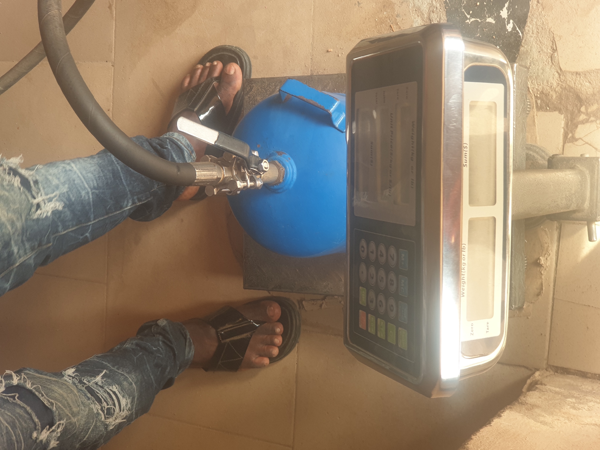In Jauro Yinu, Nigeria, this image captures a close-up view of a person's legs wearing ripped denim jeans and black sandals, standing near a digital weighing scale. A blue gas cylinder with a hose attached is placed on the scale, suggesting a weighing or refilling operation. The scale's display is visible, showing labels like "Weight(kg or lb)," "Tare," "Zero," "Unit price(s)/kg or S/bul," and "Sum($)". The environment appears to be an indoor space with a tiled floor. The lighting is somewhat uneven, with a bright glare on the scale. The image focuses on the utilitarian aspects of the scene, highlighting the scale and the gas cylinder.