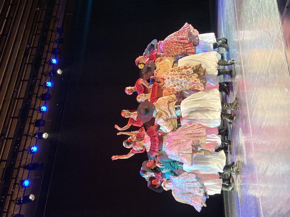 A group of approximately 15-20 individuals performs a choreographed dance on an indoor theatrical stage. The stage features a dark, unadorned background and a moderately reflective floor. Overhead, a visible grid of stage lighting fixtures includes several active blue and a few white spotlights illuminating the performers.
The individuals, a mix of male and female dancers, are clustered in a dynamic, semi-circular formation in the center of the stage. Female dancers wear traditional-style costumes consisting of ruffled, off-the-shoulder tops primarily in red or terracotta colors, paired with full, layered skirts featuring floral patterns in shades of pink, cream, and orange, or solid white. Some female dancers have adornments in their hair. Male dancers are dressed in traditional gaucho-style attire, including dark, wide-brimmed hats, loose-fitting white or cream trousers (bombachas de campo), dark boots, and various patterned or solid-colored shirts, some with sashes or belts.
Dancers are captured in various poses; several have their arms raised in expressive gestures, and many exhibit smiles. One female dancer in the foreground appears to be in mid-air or executing a jump, indicating active movement. The precise venue within the city of Buenos Aires, Argentina, cannot be determined from the visual evidence provided.