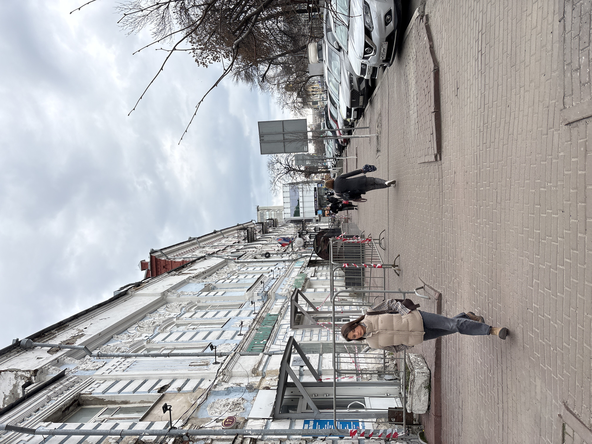 The image depicts a street scene in Ulyanovsk, Russia. An older building with peeling white paint and intricate architectural details lines the left side of the frame. The sky is overcast. A woman wearing a beige vest and dark pants stands on a brick sidewalk in the foreground. Metal barriers wrapped with red and white tape are positioned in front of the building. Further down the sidewalk, another person walks away from the camera. A row of parked white cars is visible on the right side of the street. Bare tree branches extend across the top of the image. There's a sign pole on the right sidewalk in front of the parked cars.