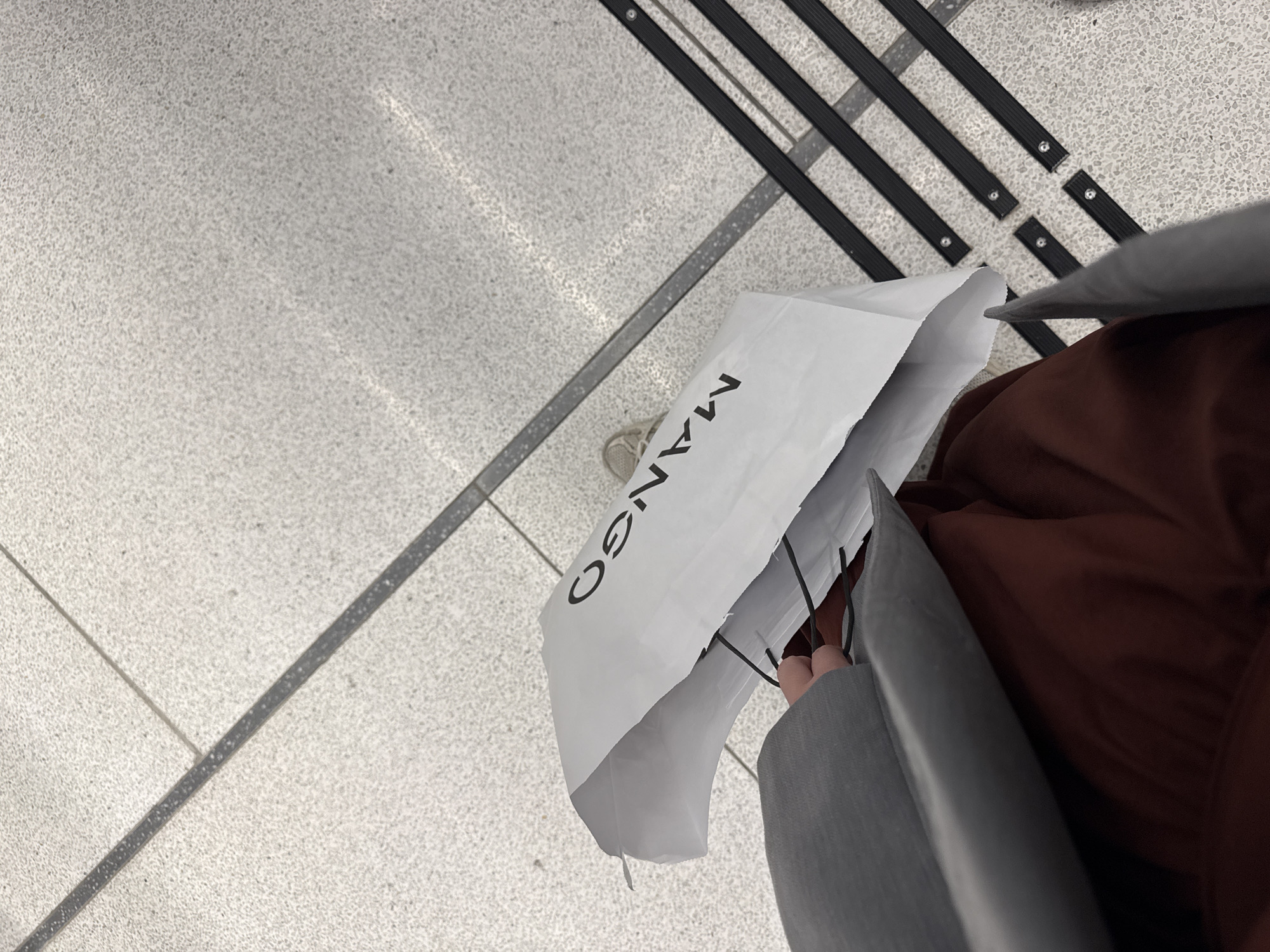 The image shows a person holding a white Mango shopping bag in a public space. The ground is covered in light gray tile, with raised black tactile paving indicators. The person is wearing a gray coat and brown clothing. The setting appears to be an indoor location, possibly a shopping mall or transit station. There is nothing in the image to suggest that it is in Tel-Aviv, Israel.