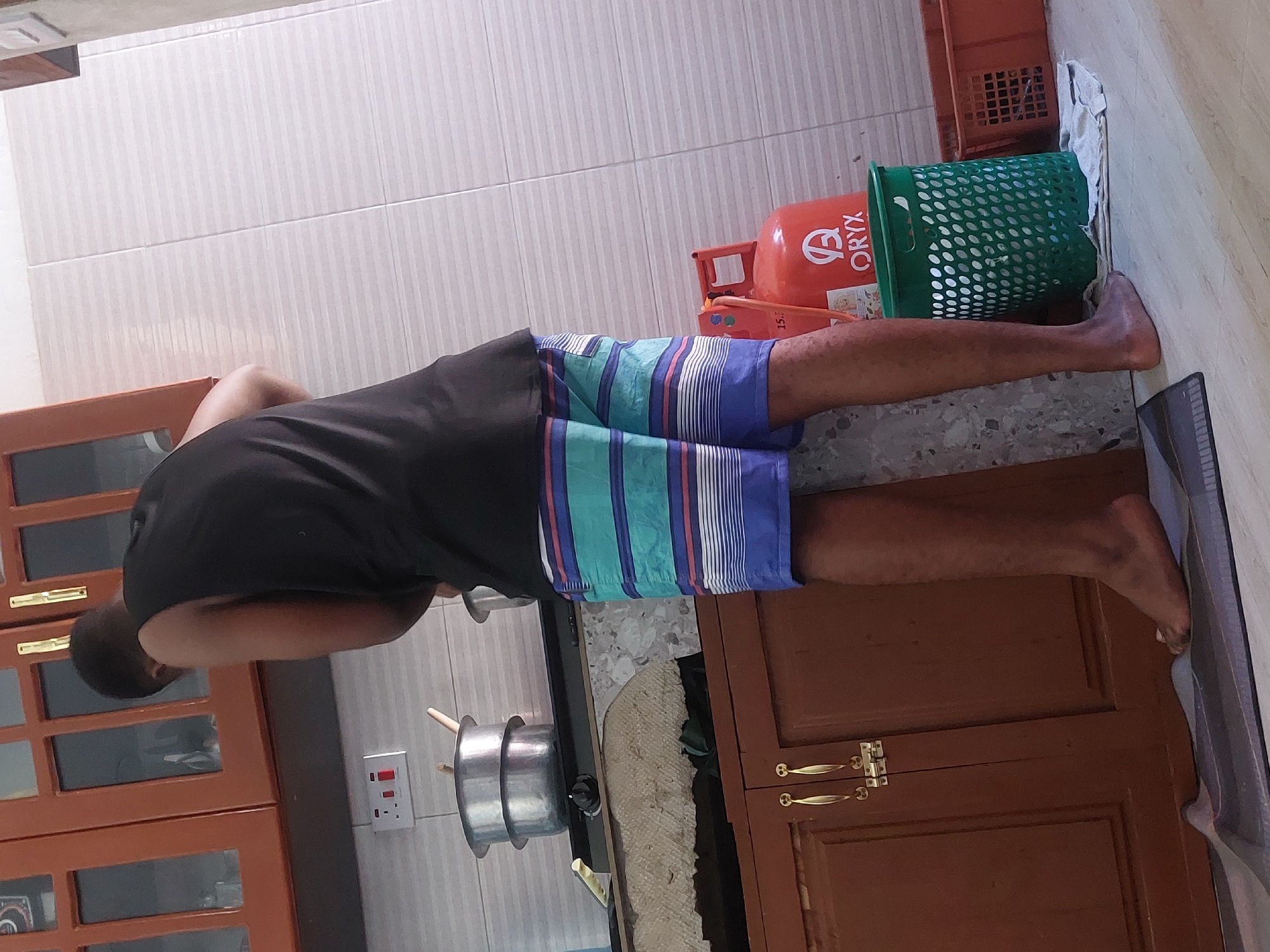 A person wearing a black shirt and blue striped shorts stands in a kitchen, next to a stovetop where a silver pot is positioned. A red Oryx gas cylinder sits near a green plastic bin, and a brown crate is partially visible in the background. The location is likely a residential kitchen in Dar es-Salaam, Tanzania, given the presence of the Oryx gas cylinder, a common brand in the region.