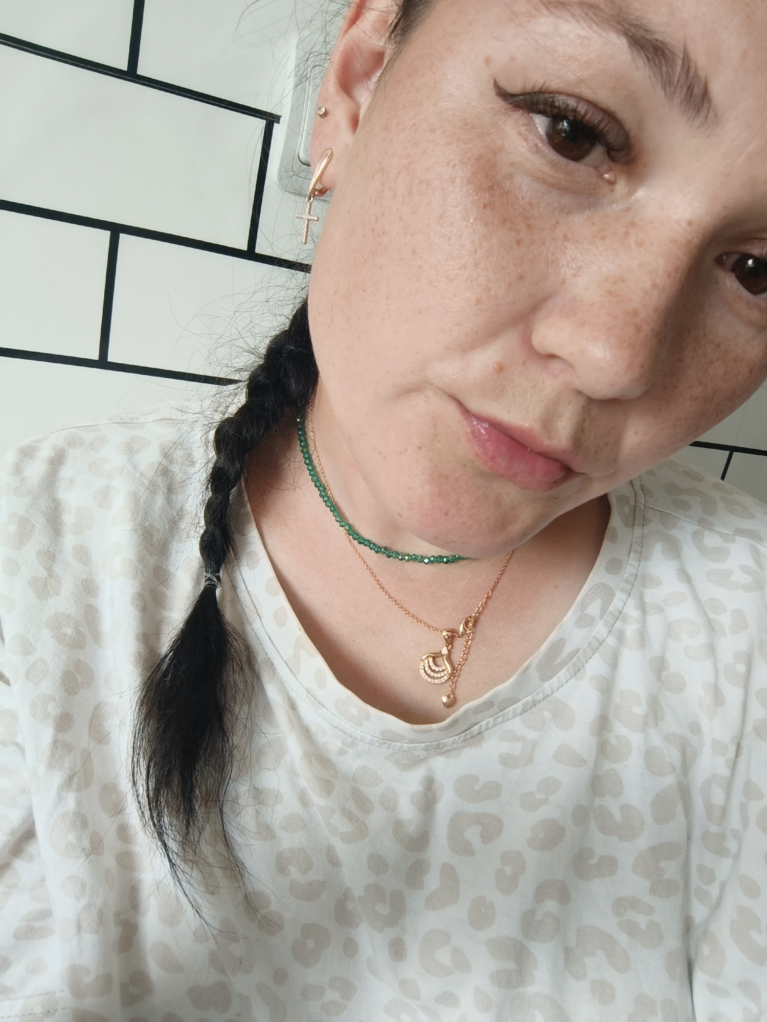 A close-up, chest-up shot shows a woman with dark hair braided and resting on her shoulder. She is wearing a white t-shirt with a light brown leopard print. She has two necklaces on; one appears to be a delicate green beaded necklace, and the other is a rose gold chain with two pendants. The woman has freckles on her face and is wearing subtle makeup, including eyeliner. She is also wearing a gold cross earring with small white stones. The background consists of a white tiled wall with black grout lines. The lighting suggests an indoor environment.