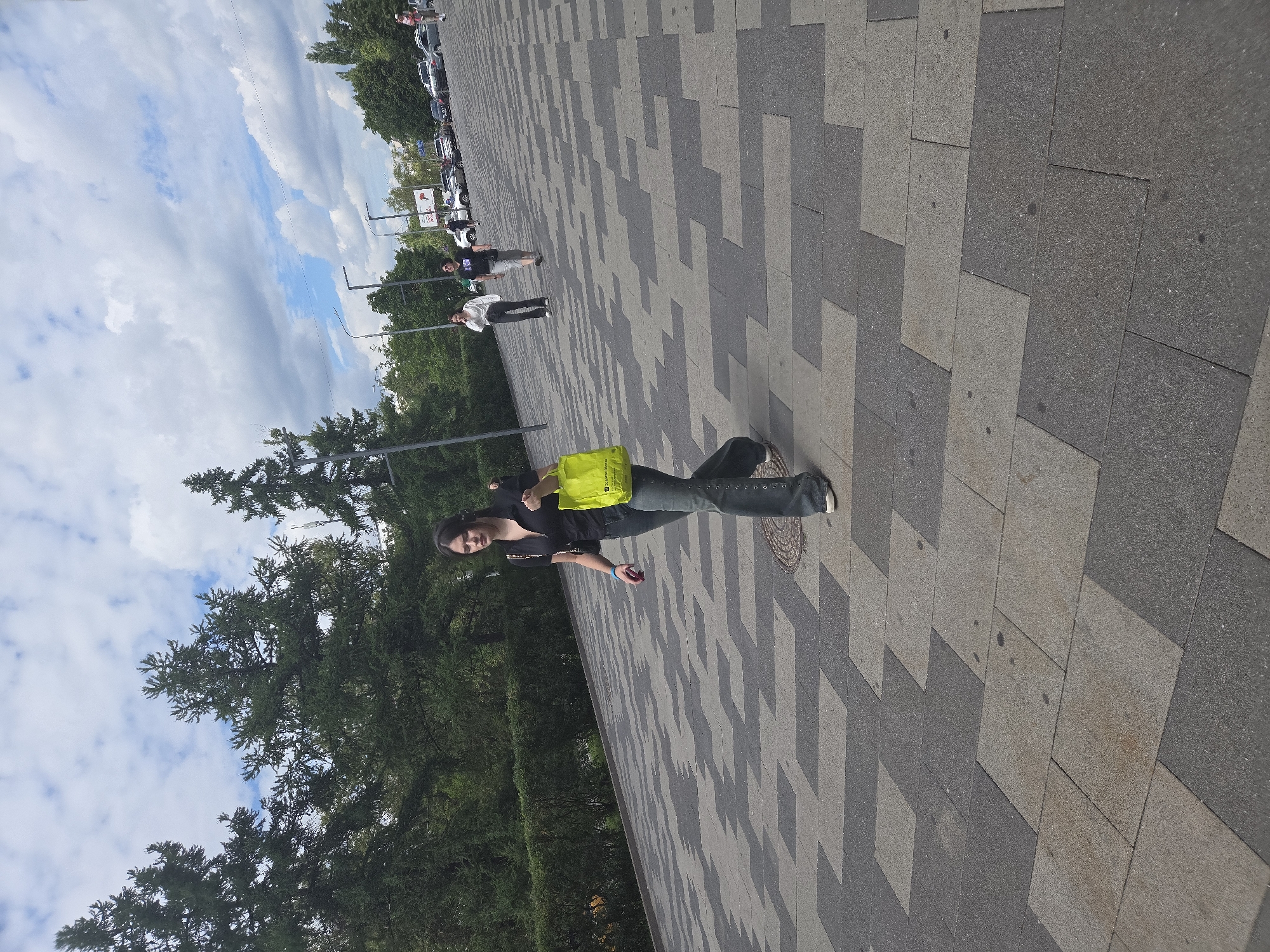A young woman walks across a paved area with a patterned stone surface. She is wearing a black shirt, gray pants, and white shoes, and she carries a bright yellow bag in her right hand. Behind her, other people are walking in the distance, and cars are parked in a lot. The background includes trees and a cloudy sky. This scene is likely in Moscow, Russia, given the style of the architecture and the overall atmosphere.
