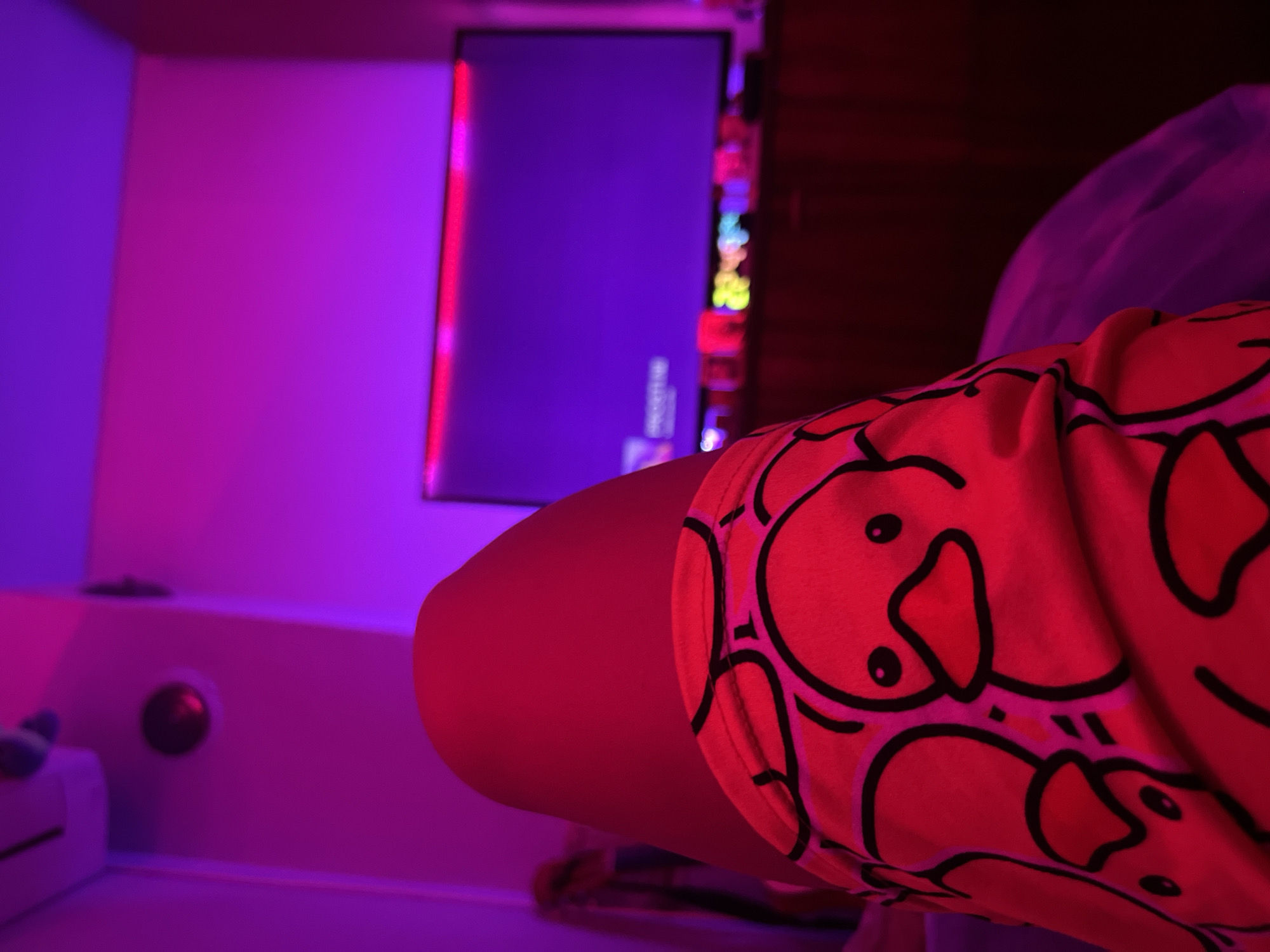 In Debelt, Bulgaria, the image presents a first-person perspective focused on the upper leg of an individual, likely seated or reclining. The leg is partially covered by a garment featuring a repeating pattern of cartoon ducks with black outlines and orange beaks on a red-orange fabric. The visible skin of the leg is smooth and appears reddish due to ambient lighting.
The background is out of focus, illuminated predominantly by intense magenta and purple light sources, casting a vibrant hue on the walls and ceiling. In the upper left quadrant, a white, horizontally oriented object, possibly an air conditioning unit or smoke detector, is mounted on the wall. Below it, a dark circular object, possibly a camera or light fixture, is also visible.
A large, dark flat-screen television is centered in the mid-background, displaying a faint purple screen with blurred white text on its left side, suggesting an active interface. Behind the top edge of the television, a horizontal strip of red LED lighting is visible. Below the television, on a dark piece of furniture, multiple small, indistinct, brightly colored, glowing objects are arranged, suggesting decorative lights or electronic devices. The overall environment appears to be an interior room, characterized by low light and specific ambient lighting.