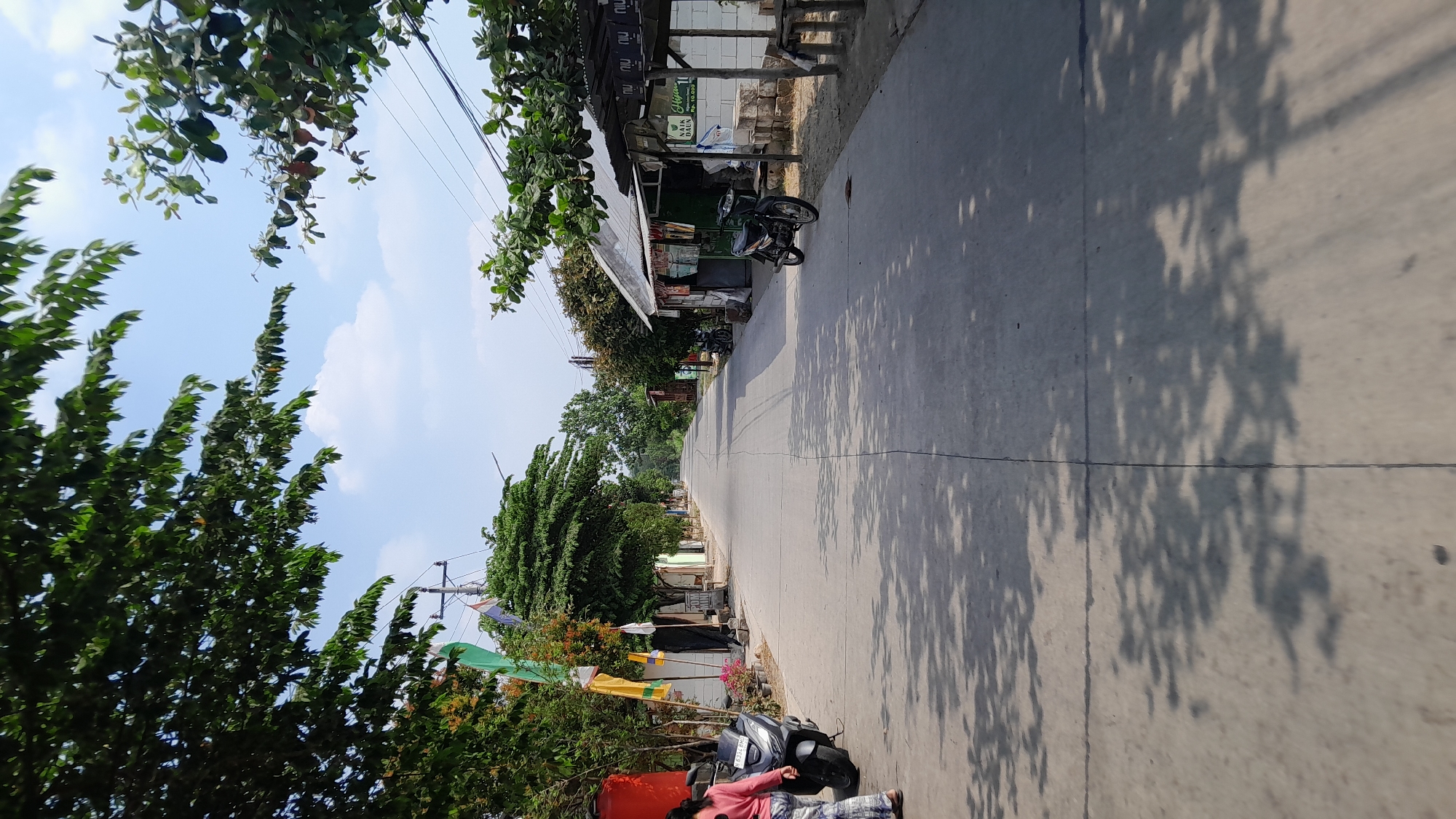 The image captures a paved road in Sumberarum, Indonesia, lined with trees and simple structures on either side. The road stretches into the distance, bordered by lush green foliage and under a clear blue sky with scattered clouds. On the left side of the road, a black scooter is parked, with a person wearing a pink long-sleeved shirt and patterned pants standing next to it. Behind the scooter, a bright red cylindrical object is visible. Further down the road on the left, there are several flags and trees. On the right side, a small open-air establishment with a tin roof displays various items for sale, and another motorcycle is parked in front of it. Building materials, including what appear to be concrete blocks, are stacked nearby. The sunlight casts distinct shadows of trees across the road, indicating a bright, daytime scene. The overall impression is of a quiet, rural or suburban street in Indonesia.