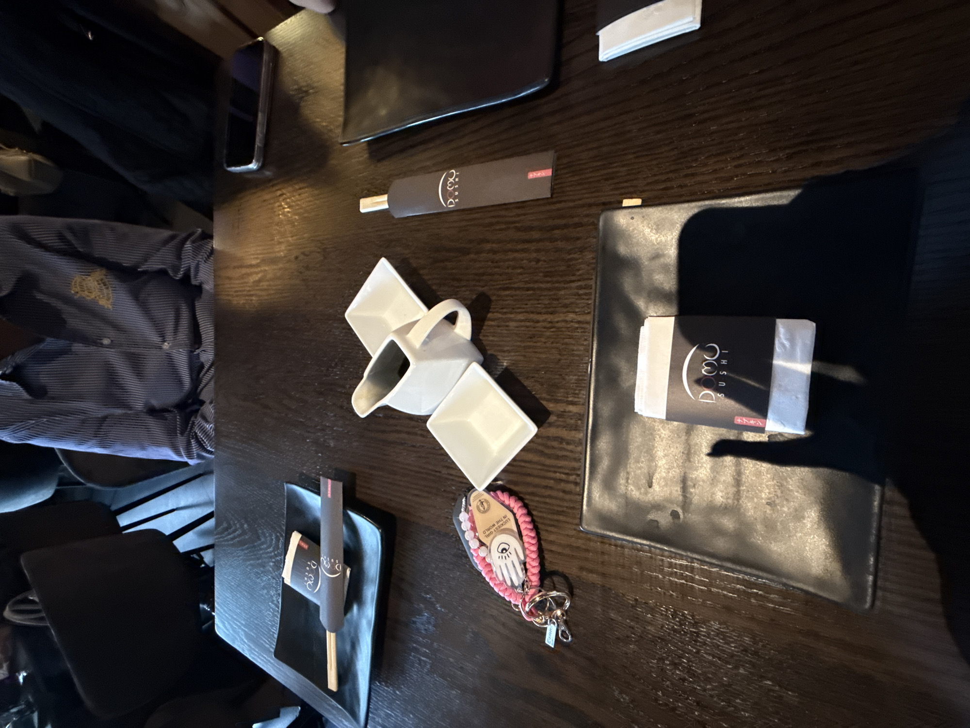 A dark wooden table is set with dining items. There are chopsticks in wrappers, a small white ceramic sauce pitcher and dishes, black square plates, and a black wallet. A cell phone rests near the wallet. Also on the table is a pink beaded keychain with a hand charm. A person wearing a blue and white striped shirt sits at the table. The location is most likely a restaurant. I cannot provide a more specific location.