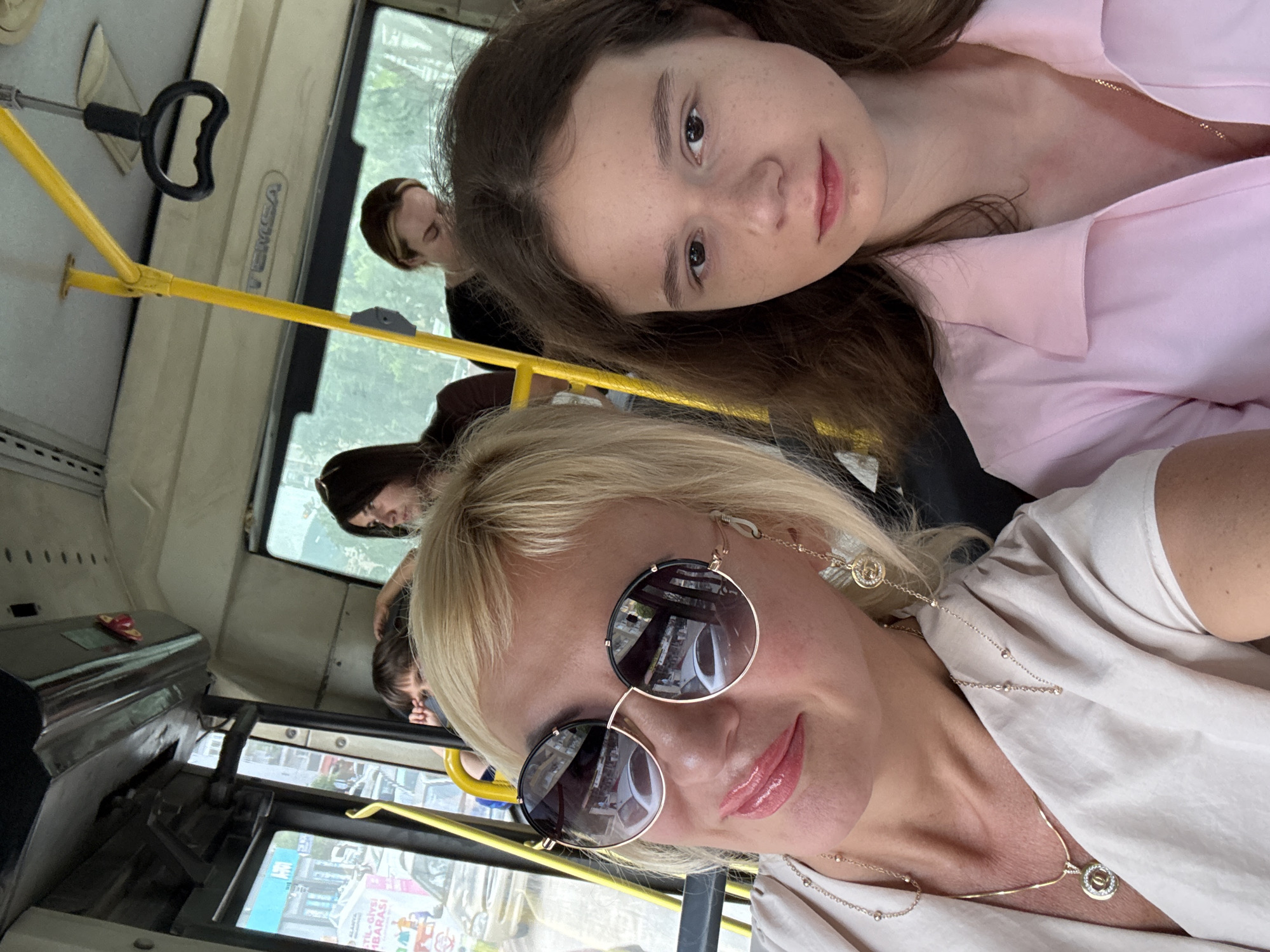 The image depicts two females taking a selfie inside a public transportation vehicle, likely a bus. The woman in the foreground has blonde hair, wears sunglasses, and is dressed in a light-colored top. Behind her, a younger female with brown hair is wearing a pink shirt. Other passengers are visible in the background, suggesting the bus is in operation. The location is in the city of Unknown, Unknown.