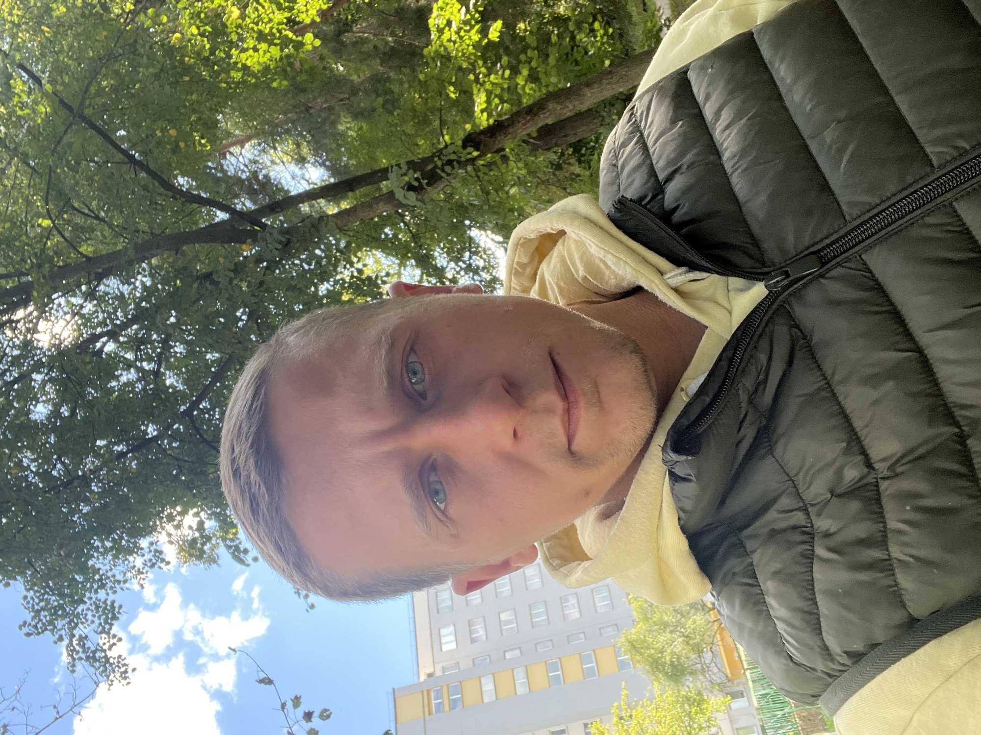The image is a close-up selfie of a man with short, light brown hair and blue eyes, wearing a light yellow hooded sweatshirt underneath a dark quilted vest. He is looking directly at the camera. In the background, to the left, is a tall, light-colored apartment building with numerous windows, characteristic of Soviet-era architecture found in Minsk, Belarus. To the right and behind the man, there are trees with green leaves, suggesting an outdoor, possibly park-like setting within the city. The sky is visible with scattered white clouds and sunlight filtering through the trees.