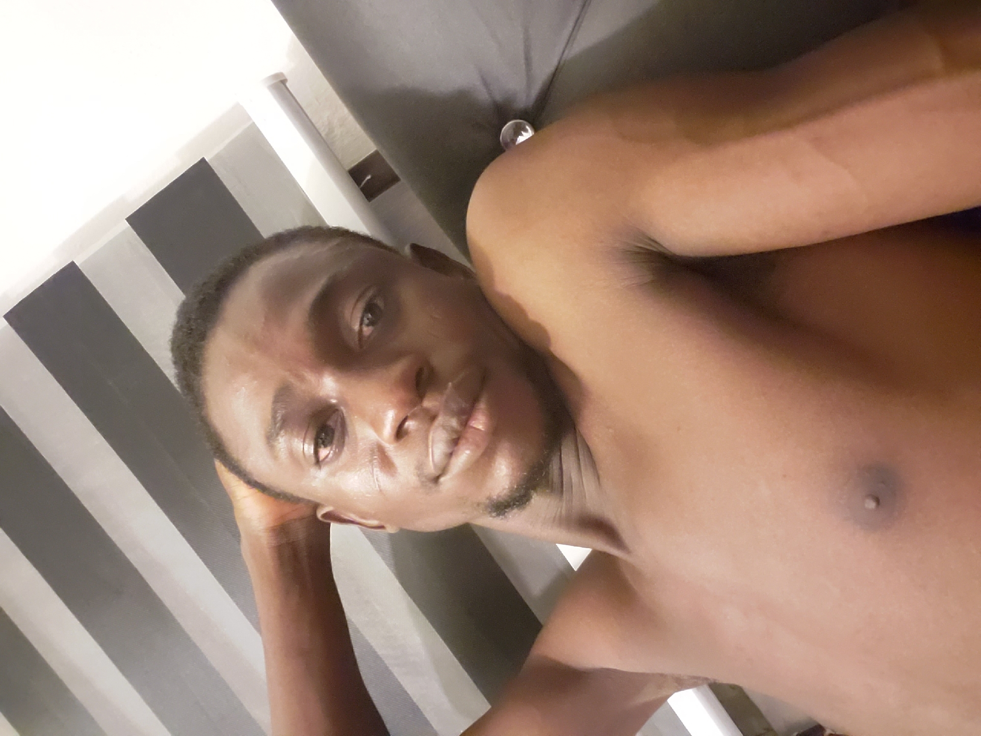 The image is a slightly tilted, close-up selfie of a young man, likely in his 20s or 30s, reclining indoors. He has dark skin, short dark hair, and a short goatee. He is shirtless, revealing his chest and shoulder. His left arm is bent, with his hand resting near his head, supporting himself as he looks directly into the camera with a neutral expression. His skin appears to have a slight sheen, possibly from sweat or the indoor lighting.
The background on the left features a wall with alternating vertical black and light grey stripes. On the right, behind his head and right shoulder, is a dark grey or black surface, likely a bed or sofa. A clear, spherical object is visible on this dark surface near his right shoulder. A small, light-colored nipple piercing is visible on his chest. The lighting is consistent with an indoor setting, and no specific time of day can be determined. There is no visible text. This scene is likely set within a private residence in Adonkolo, Nigeria.