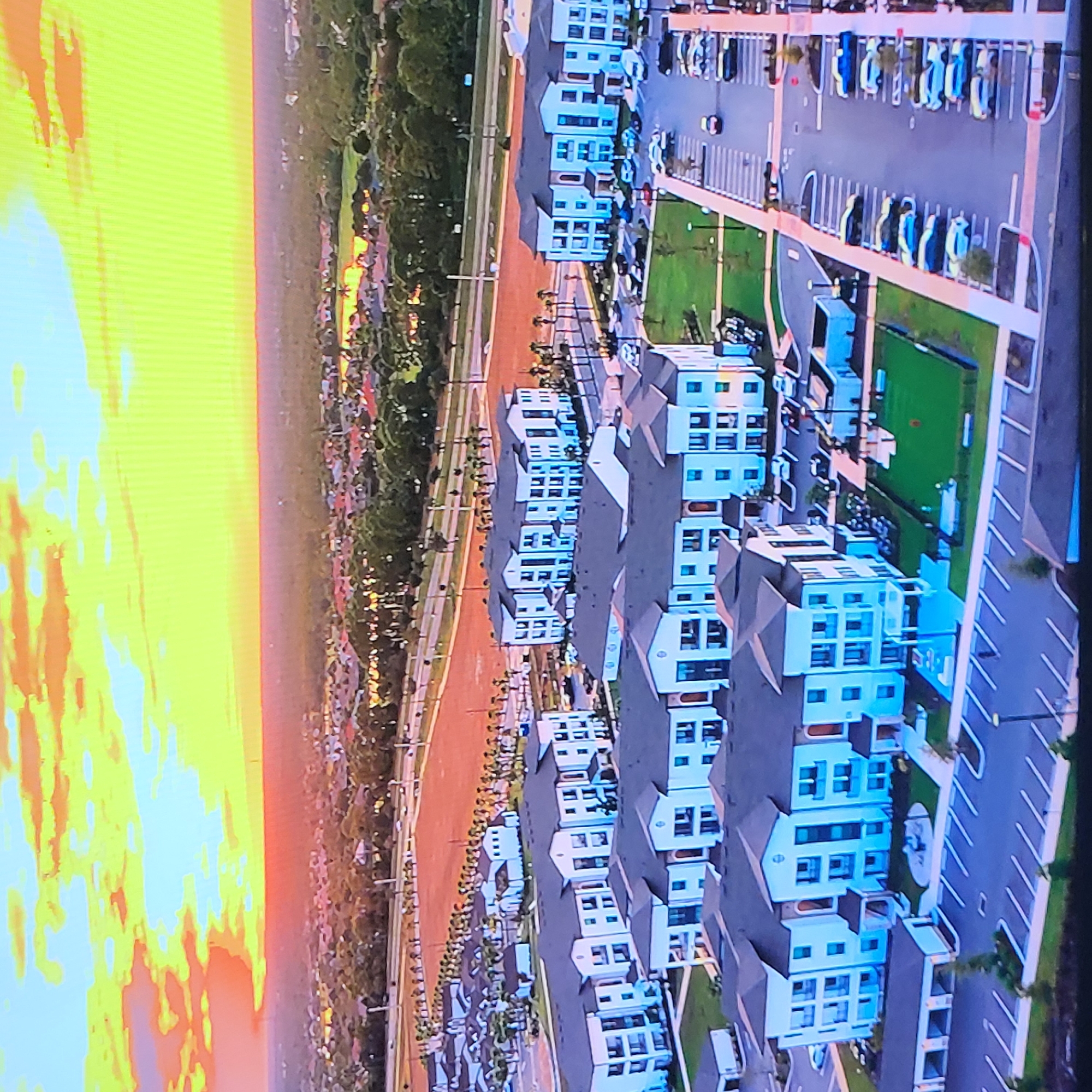 The image is an aerial view of a residential neighborhood. There are multiple buildings with a similar architectural style, most appearing to be three stories tall, and painted white with dark roofs. A road runs parallel to the buildings, with trees planted along it. There is a parking lot with many parked cars. In the distance, there is a band of greenery that extends across the frame, suggesting trees or parkland. To the left, there is a bright sky with hues of yellow, orange, and blue, resembling a sunset or sunrise. The city of Yerevan, Armenia, is not depicted or recognizable in this image.
