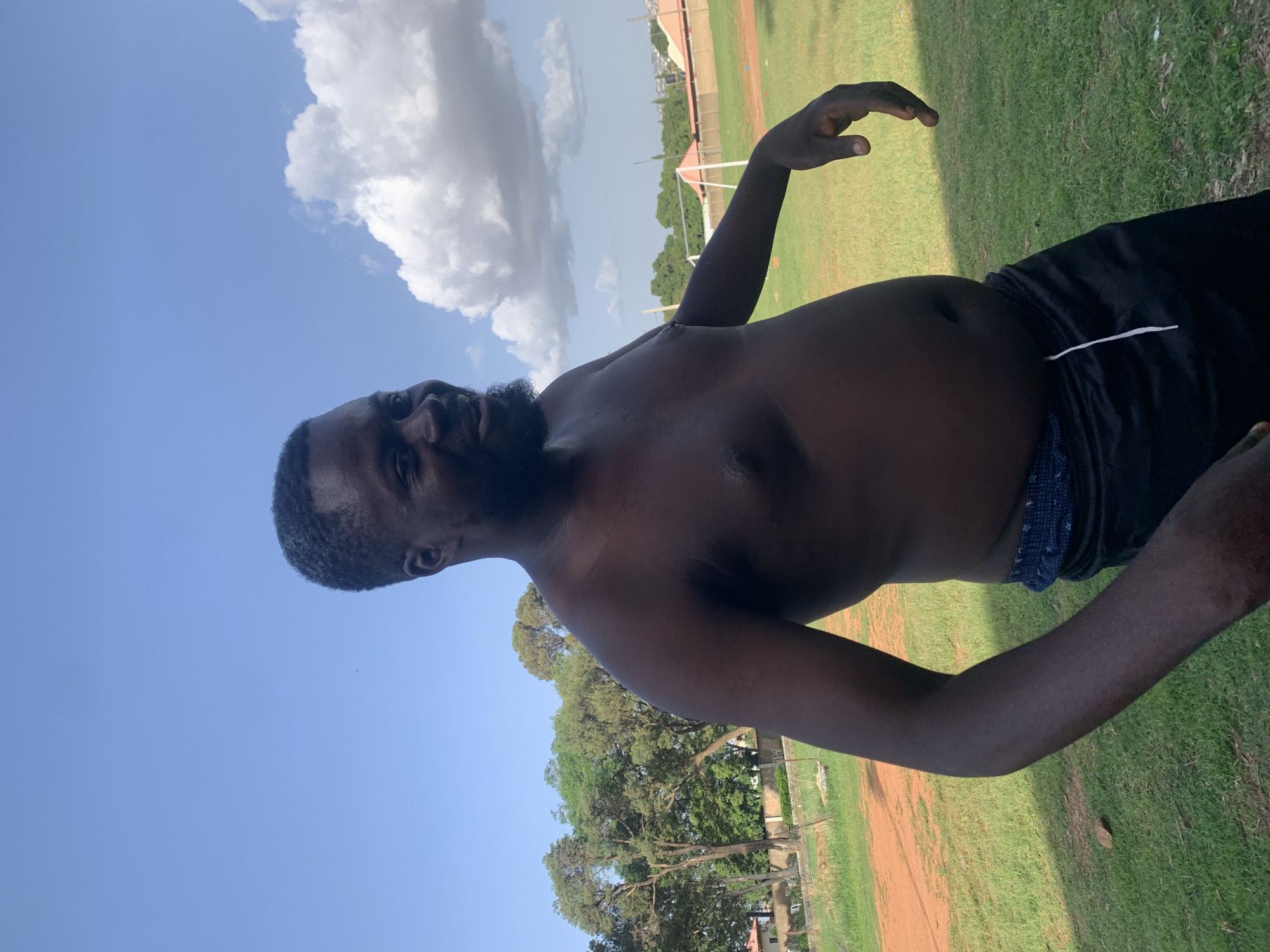 A shirtless Black man with dark skin, a beard, and short hair is standing in a grassy field with a red dirt area to his left. He is wearing black athletic shorts with a blue patterned waistband and a white drawstring. The man is smiling and looking to his right, with his left arm bent at the elbow and his hand open. Behind him, there are trees and a cloudy blue sky. In the distance, there is a soccer goal and some buildings. The scene appears to be outdoors in a relatively open area, possibly a park or sports field, suggesting a location within the city of Jos, Nigeria, given the architectural style of the buildings in the background and the type of vegetation.
