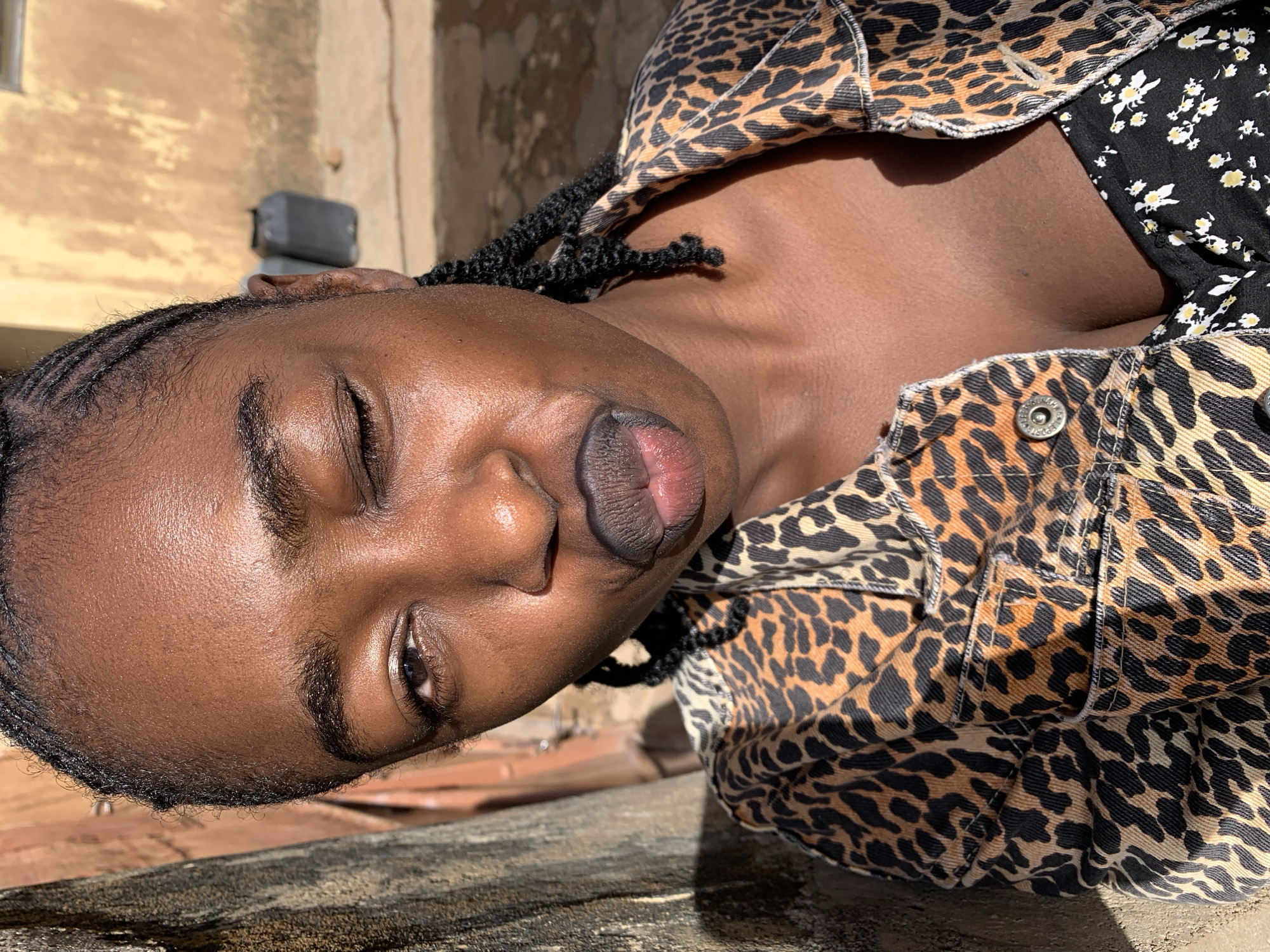 A close-up portrait of a person with dark skin, oriented such that their head is tilted to the right. The subject appears to be lying down or leaning against a surface, looking towards the viewer with their eyes partially closed or squinting in the bright sunlight, suggesting a relaxed or contemplative expression. They have braided hair and full, slightly parted lips with a reddish tint. The person is wearing a jacket adorned with a distinctive leopard print pattern, under which a black garment with a white and yellow floral print is visible around the collarbone. Intense, direct sunlight illuminates the scene, casting strong shadows on the person's neck and chest, indicating daytime. The background features a rustic, light-colored building or wall, possibly made of sun-baked earth or painted plaster, with a dark rectangular object (possibly a vent or air conditioning unit) mounted on it. The surface supporting the person is rough and appears to be made of concrete or stone. The setting is Rafin Par, Nigeria. There is no visible text in the image.