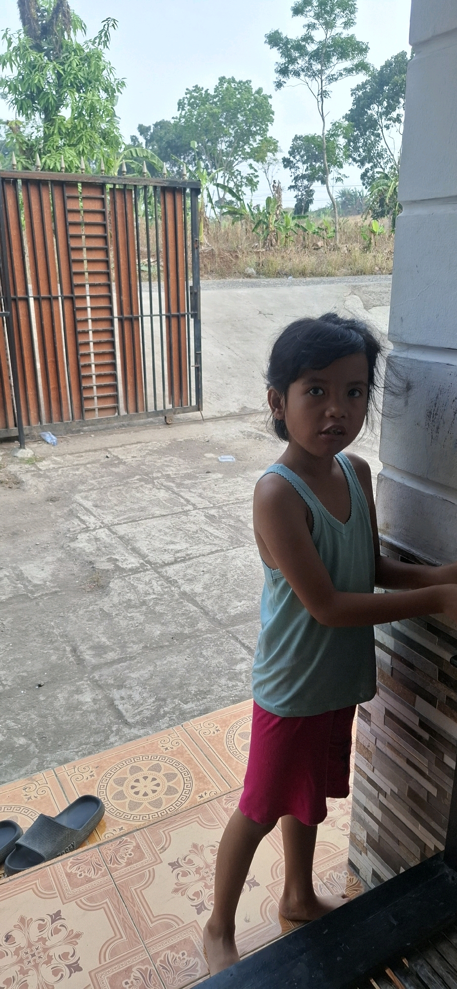 The full-body, vertical shot captures a young girl standing near a building entrance in Cibinong Hilir, Indonesia. She is positioned on a tiled porch, with a concrete walkway and a metal gate visible in the background. The girl wears a light blue sleeveless tank top and pink shorts. She stands facing the camera, looking slightly to her left, with her hands near her sides. Her dark hair is short and slightly disheveled. The porch tiles feature an ornate pattern in shades of brown and cream. To the left of the girl, on the concrete walkway, a pair of dark gray flip-flops are placed. Beyond the concrete walkway, a brown metal gate with vertical slats is open, revealing a dry, grassy area with scattered trees and vegetation under a bright, overcast sky. The building to the right of the girl has a wall with a textured surface of stacked rectangular tiles in varying earthy tones.