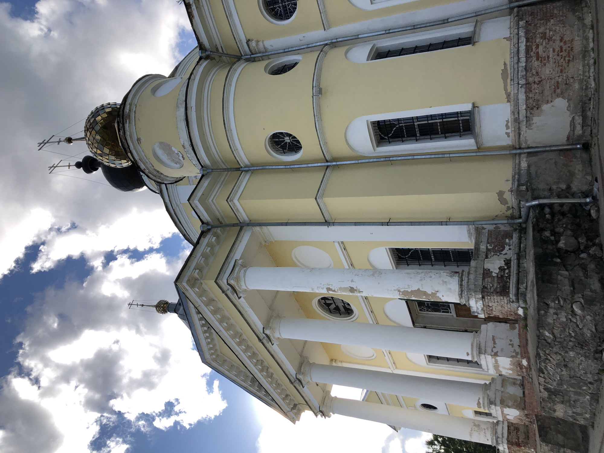 The image shows a yellow Orthodox church with white columns in the city of Myshkin, Russia. The church has several domes, one of which is golden and covered in scales. The building appears to be old, with some signs of wear and tear on the facade and columns. The sky is blue with scattered clouds. There are no people visible in the image. The architectural style suggests it's a historical building, likely a church or cathedral, typical of Russian religious architecture. The surrounding environment is not clearly depicted beyond the immediate vicinity of the church.