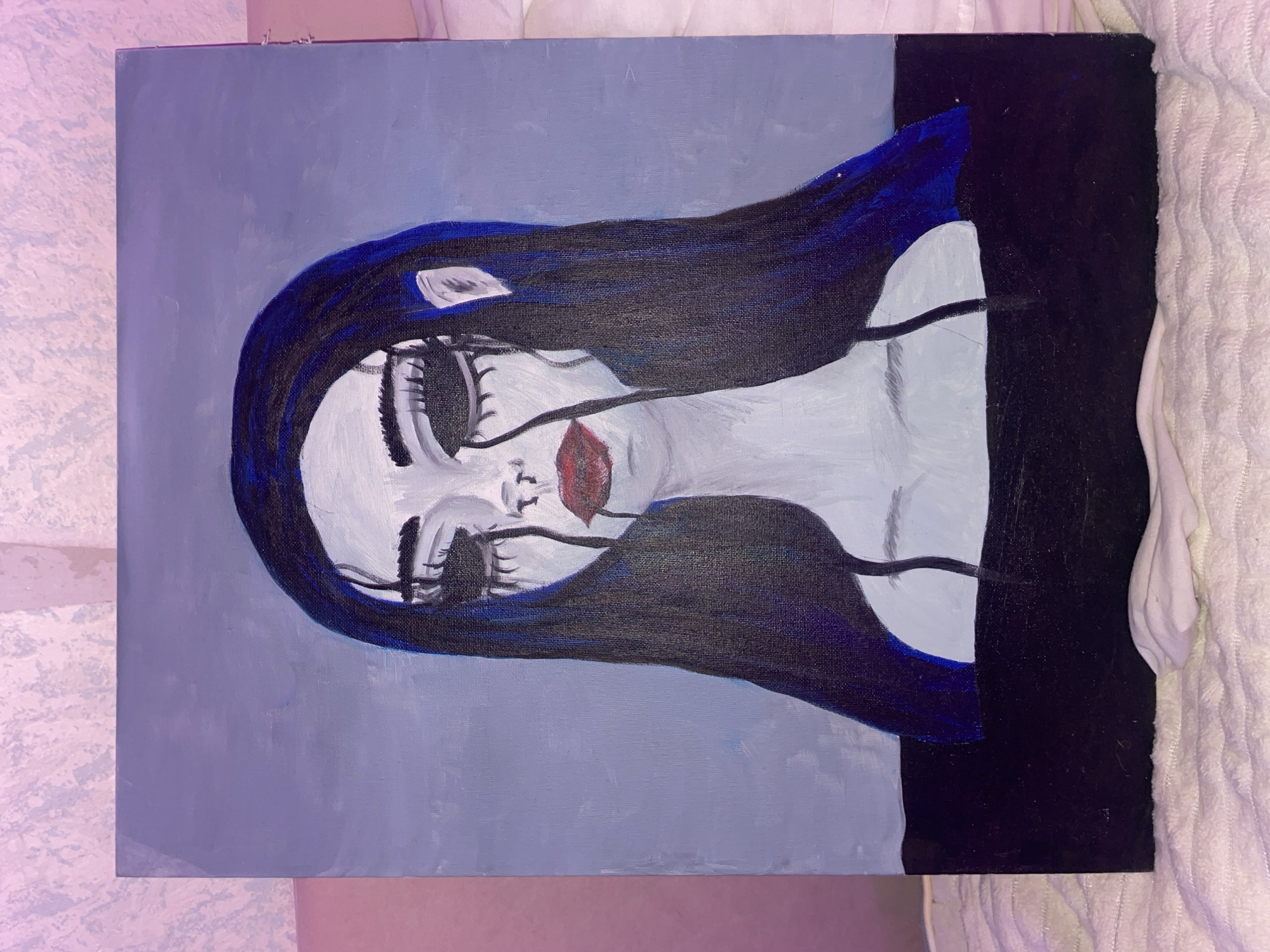 The image displays a stylized painting featuring a melancholic female figure, rendered in a somewhat surreal and gothic aesthetic. The figure's face and visible upper torso are painted in a stark, pale white tone. Her eyes are large and heavily outlined in black, with prominent, dark, streaky lines extending downwards from them, resembling exaggerated tears or melting makeup. Her lips are a vibrant red and slightly parted. Long, dark hair, with hints of deep blue, frames her face and flows down her shoulders. A small, white, ear-like shape is visible on the left side of her head.
Two thin, dark lines extend from near her shoulders down across her chest, possibly representing delicate straps or an abstract design. The figure wears a dark, possibly black, garment covering her lower torso, which blends into the lower portion of the painting's background. The background is divided into two main sections: an upper part in a muted, light grey-blue color, and a lower part in a solid dark blue to black, matching her clothing.
The painting appears to be done with acrylic or oil paints on canvas, with visible brushstrokes, particularly in the hair and background. The overall mood conveyed is one of sadness, introspection, or a doll-like fragility. There is no explicit activity taking place, as it is a static portrait. No text is visible in the painting.
The photograph itself, taken in Votkinsk, Russia, shows the painting leaning against an unseen surface, with a textured, light-colored wall visible to the left and a white, fluffy blanket or towel on the right. The ambient lighting has a subtle pinkish hue.